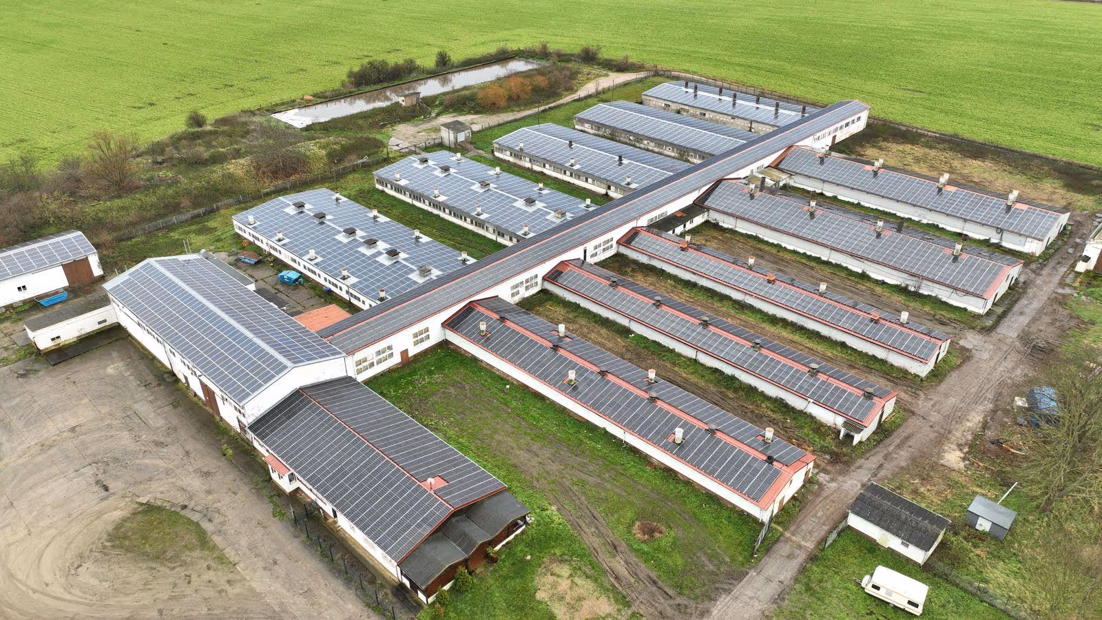 Luftaufnahme einer großen Anlage mit mehreren Gebäuden, die alle mit Solarpaneelen auf den Dächern bedeckt sind, umgeben von grünen Feldern.