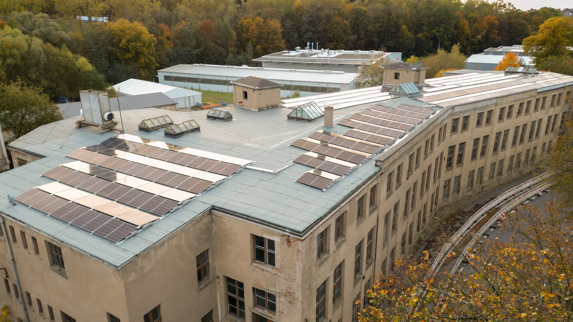 Dach eines großen Gebäudes mit mehreren Solarpanel-Anlagen und umliegenden herbstlichen Bäumen.