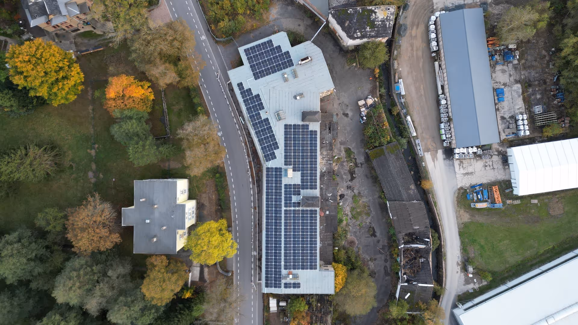 Luftaufnahme eines langen Gebäudes mit Solarmodulen auf dem Dach neben einer kurvigen Straße und Bäumen im Herbst.