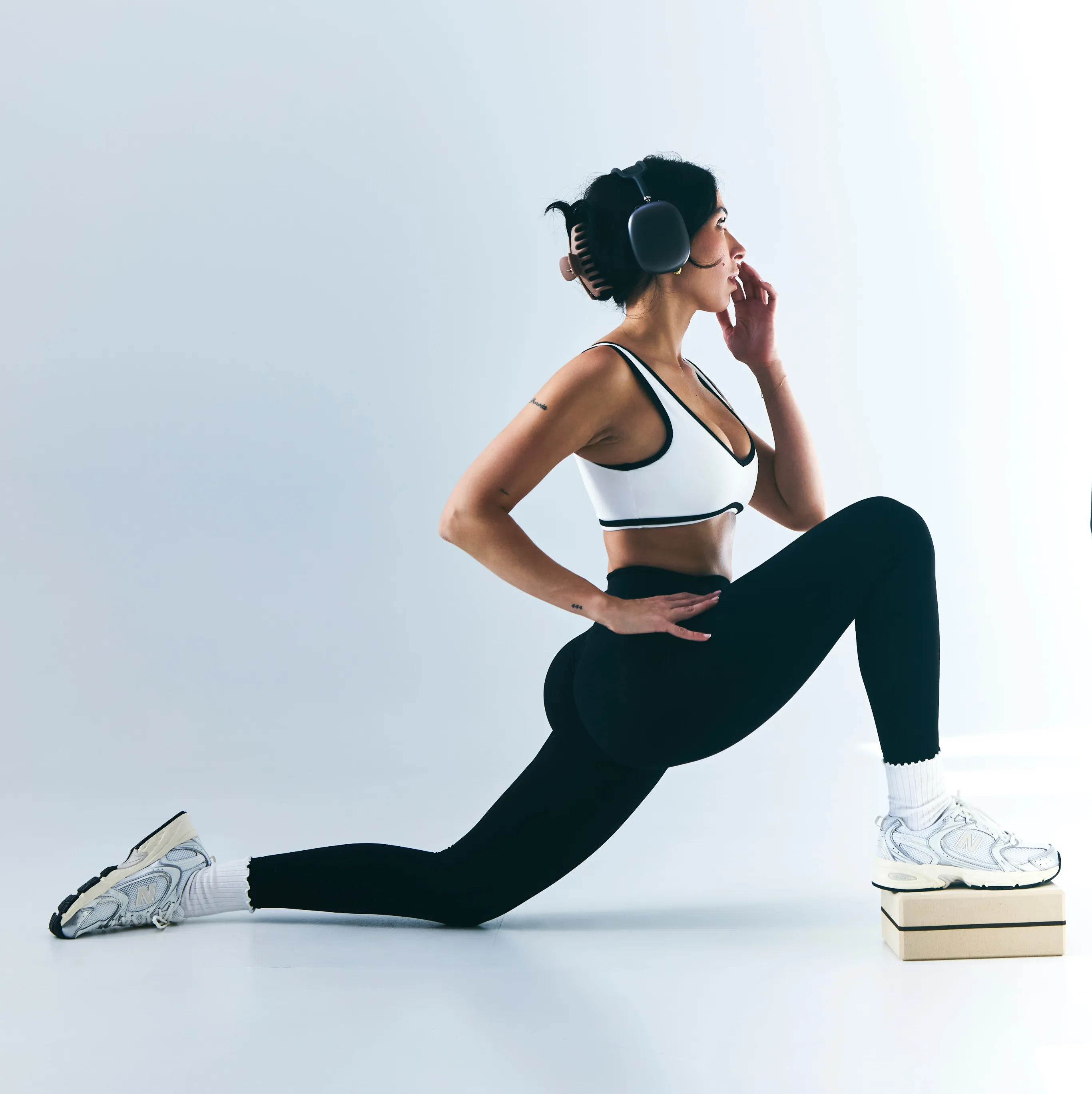 Woman in black leggings and white sports bra stretching her leg on a stack of boxes while wearing headphones.