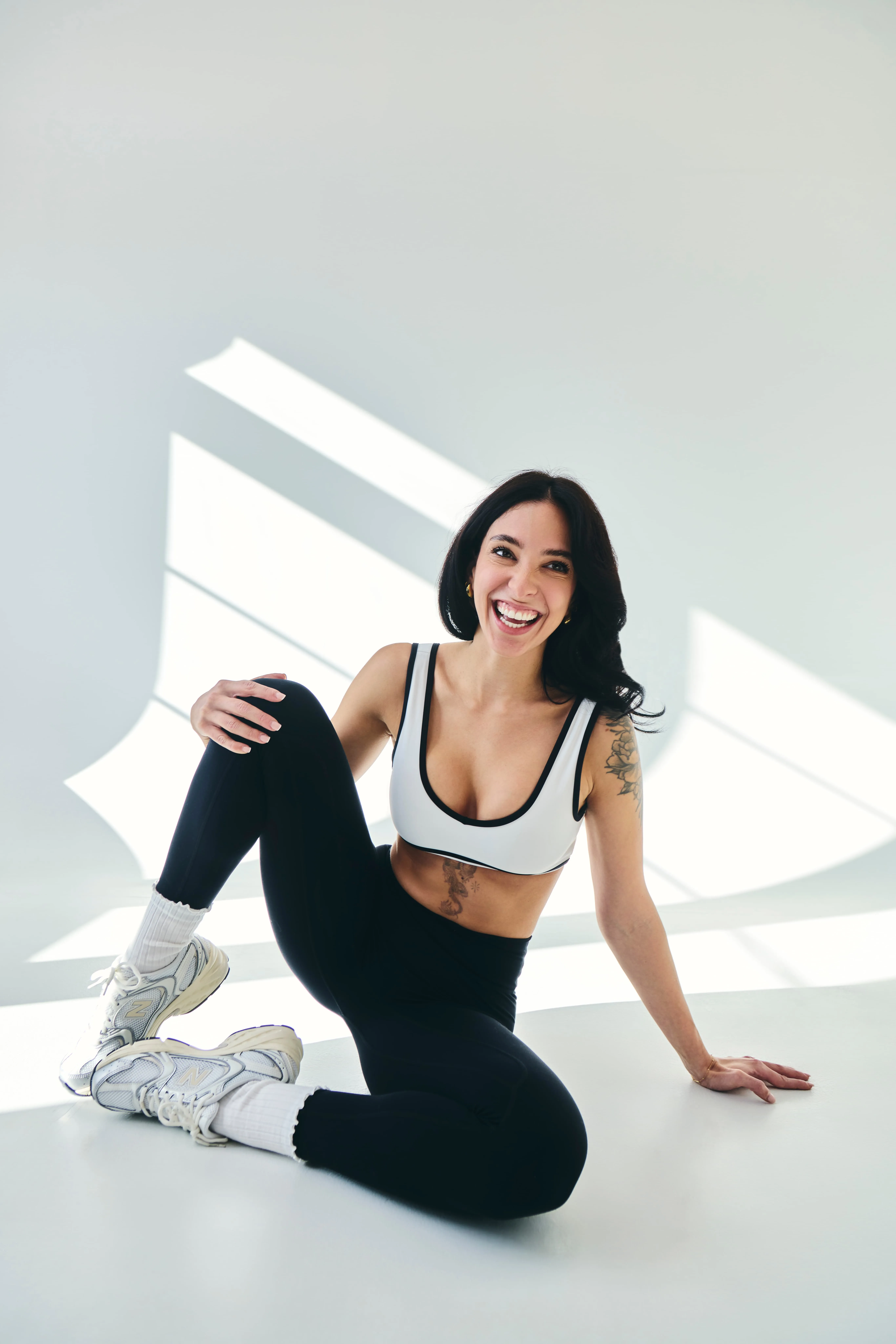 Smiling woman in black leggings and white sports bra sitting on the floor in bright room with sunlight shadows.