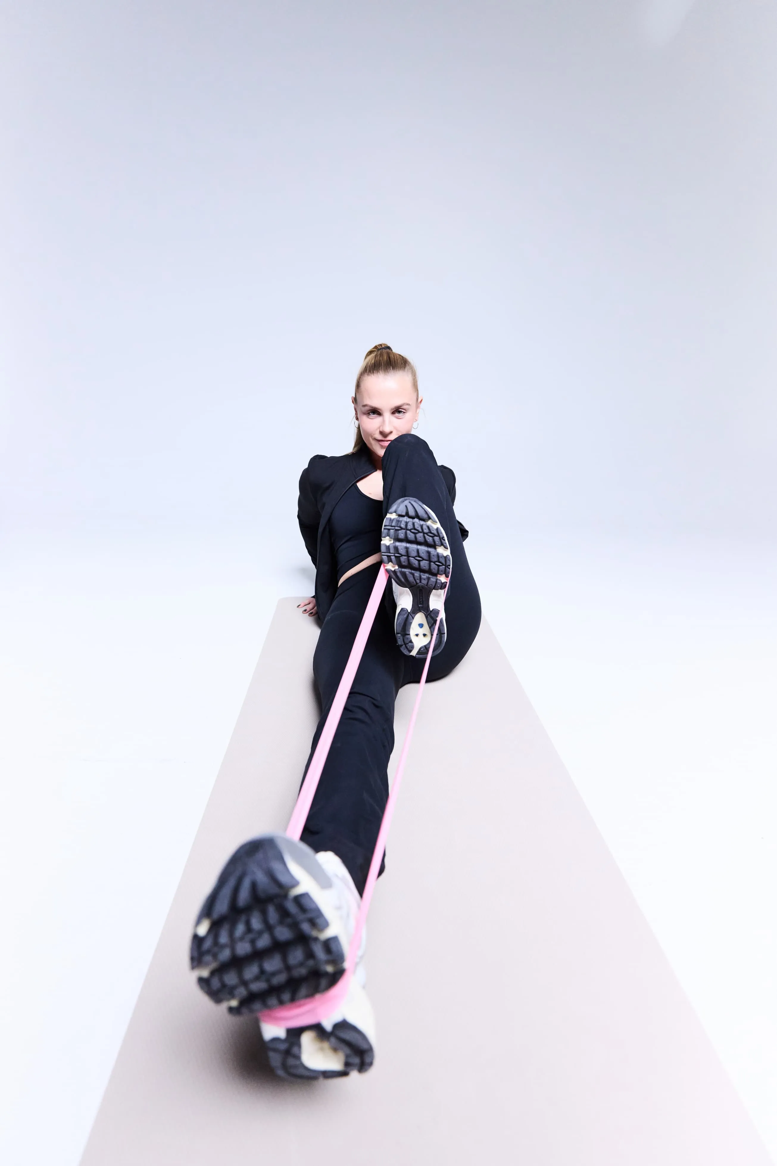 Woman in black workout clothes using a pink resistance band on her feet while lying on a light gray exercise mat.