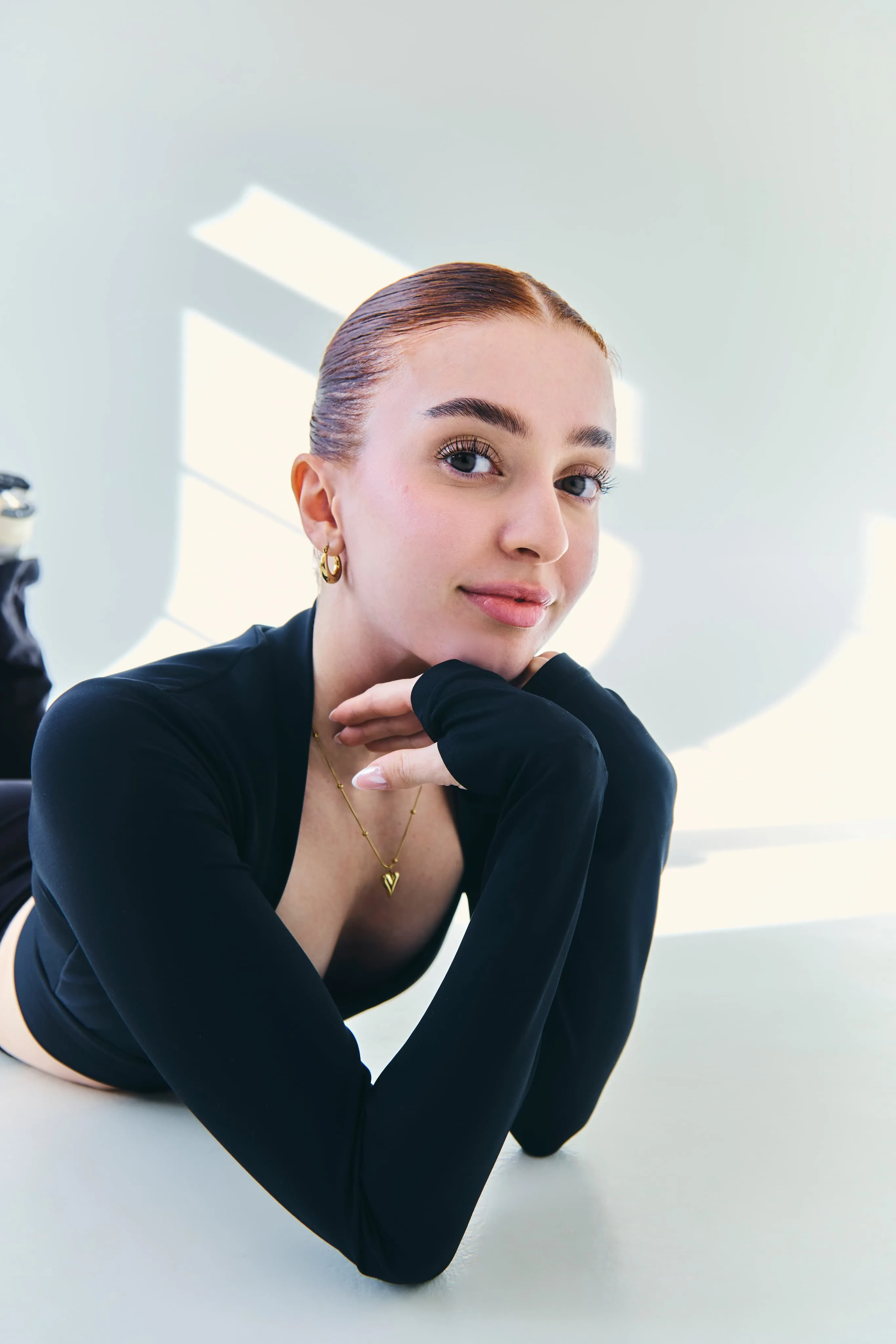 Young woman with slicked-back hair resting her chin on her hands, wearing a black long-sleeve top and gold jewelry in a bright room.