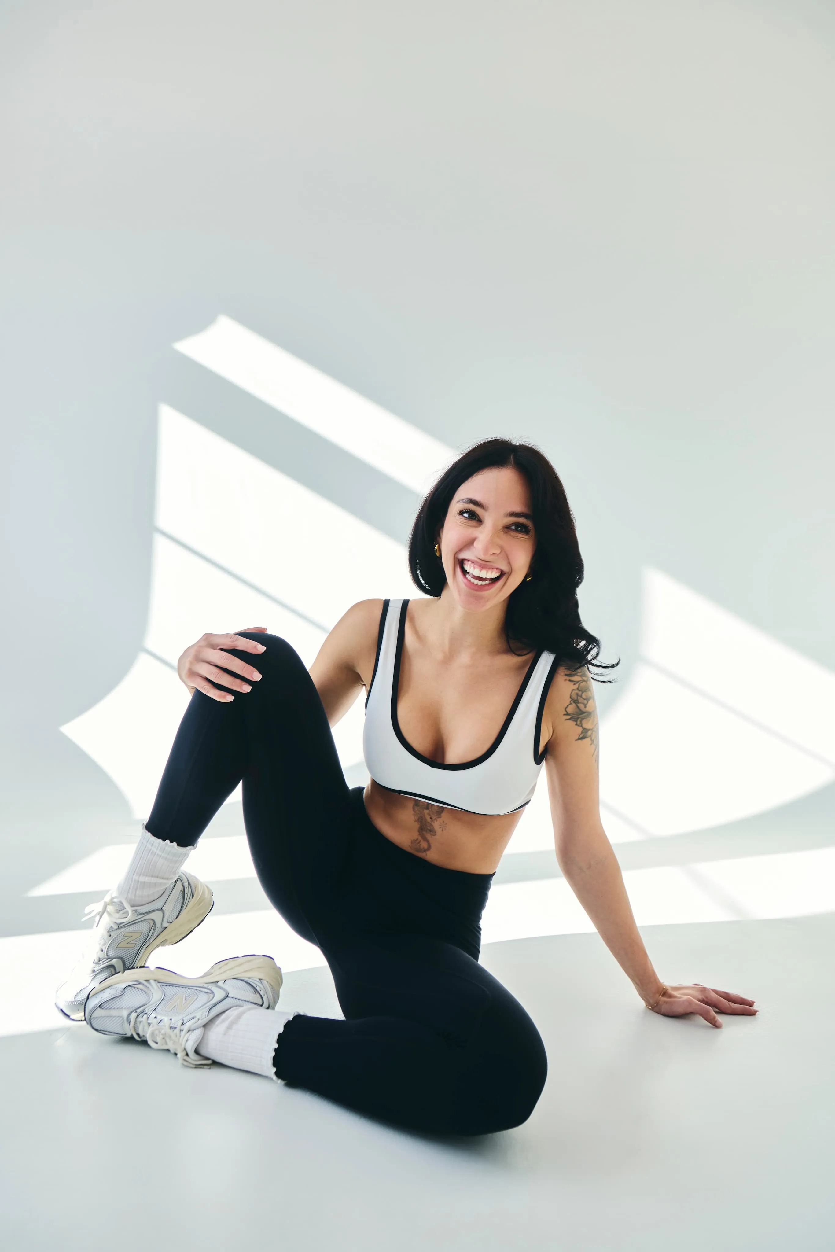 Smiling woman in sportswear sitting on the floor in a bright room with geometric light patterns behind her.