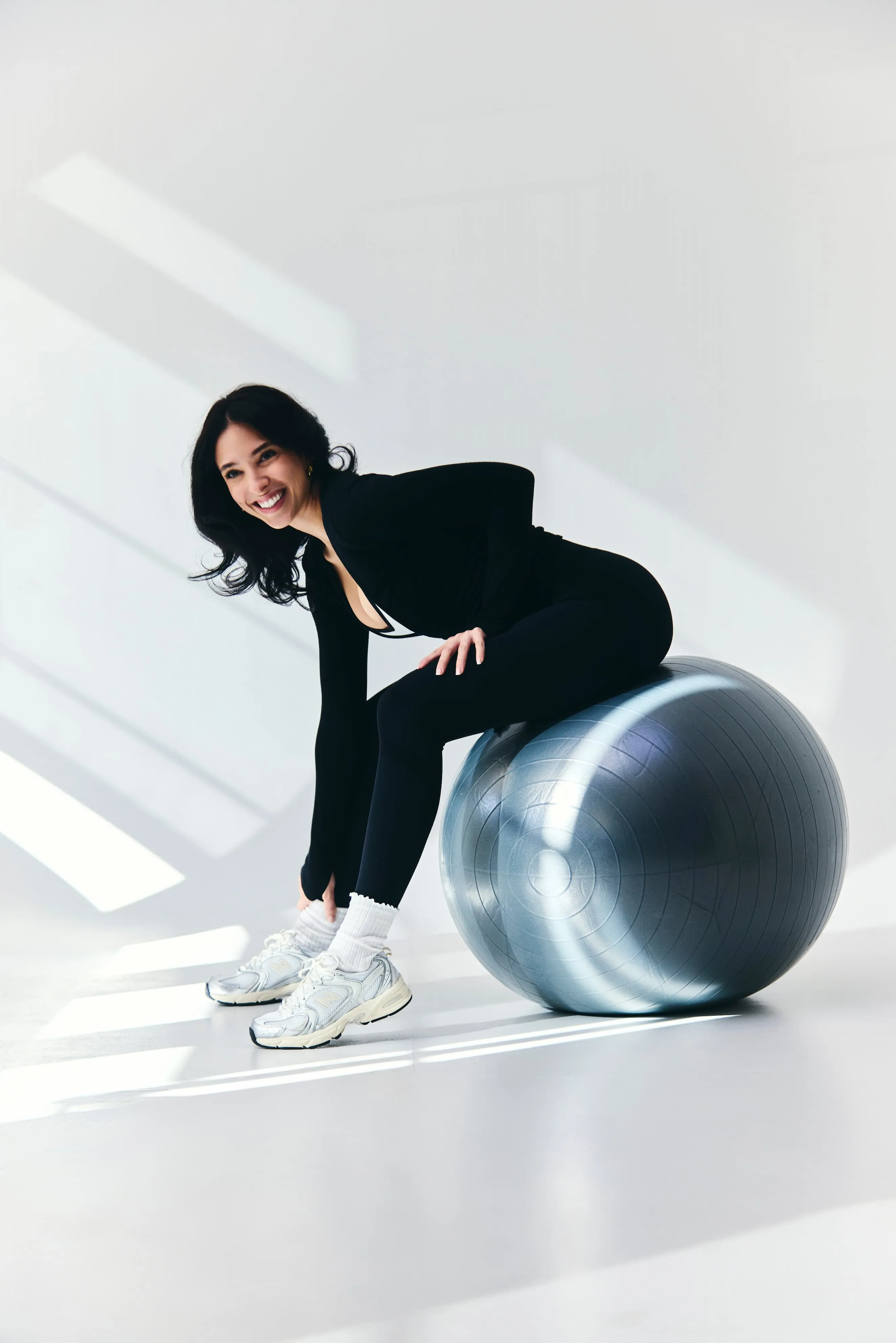 Smiling woman in black athletic wear sitting on a silver exercise ball in a bright studio.
