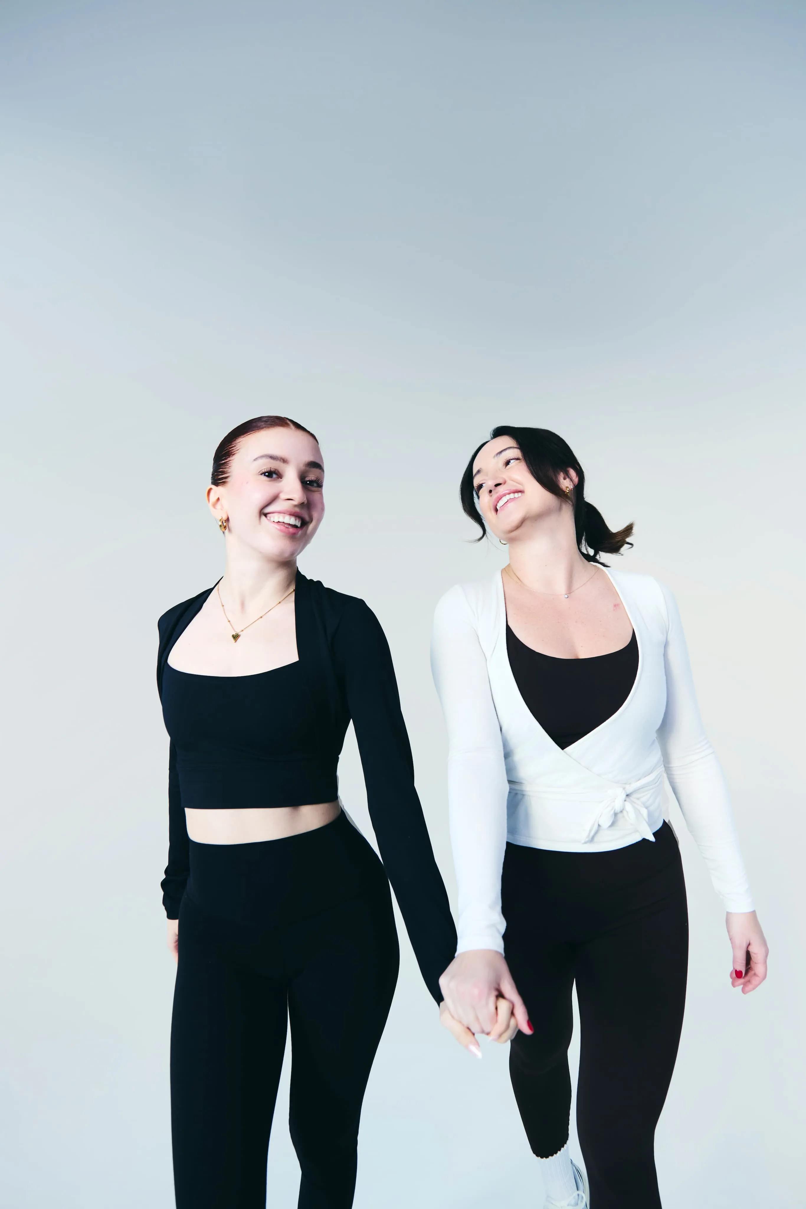 Two young women holding hands, smiling, and walking against a light gray background.
