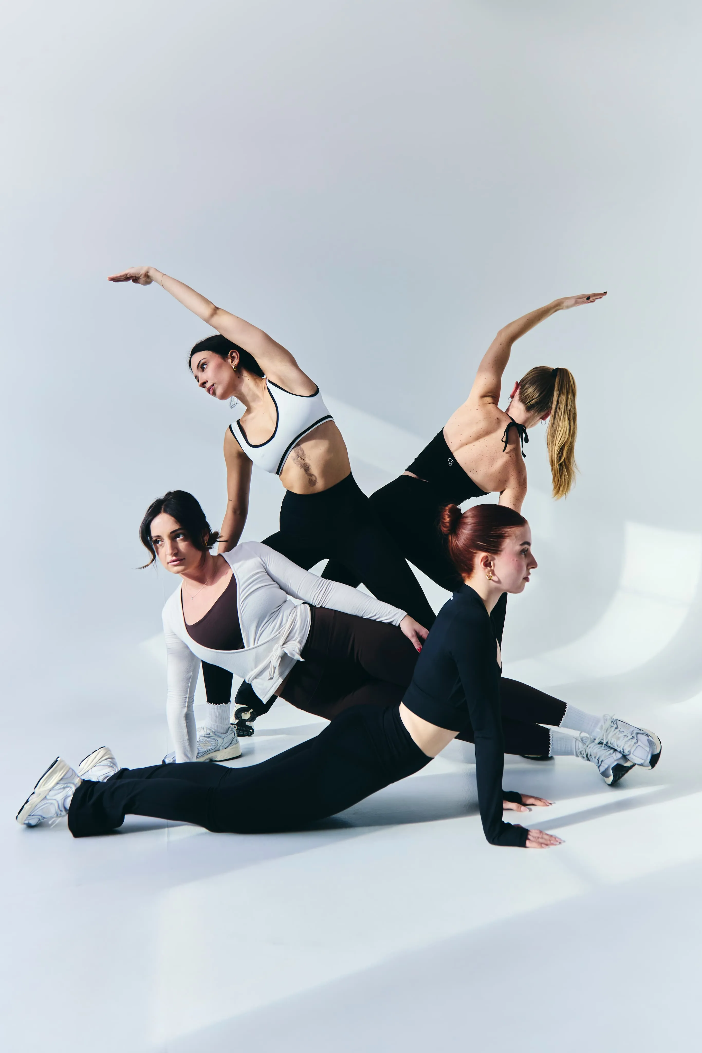 Four women in athletic wear stretching and posing against a white background with soft natural light.