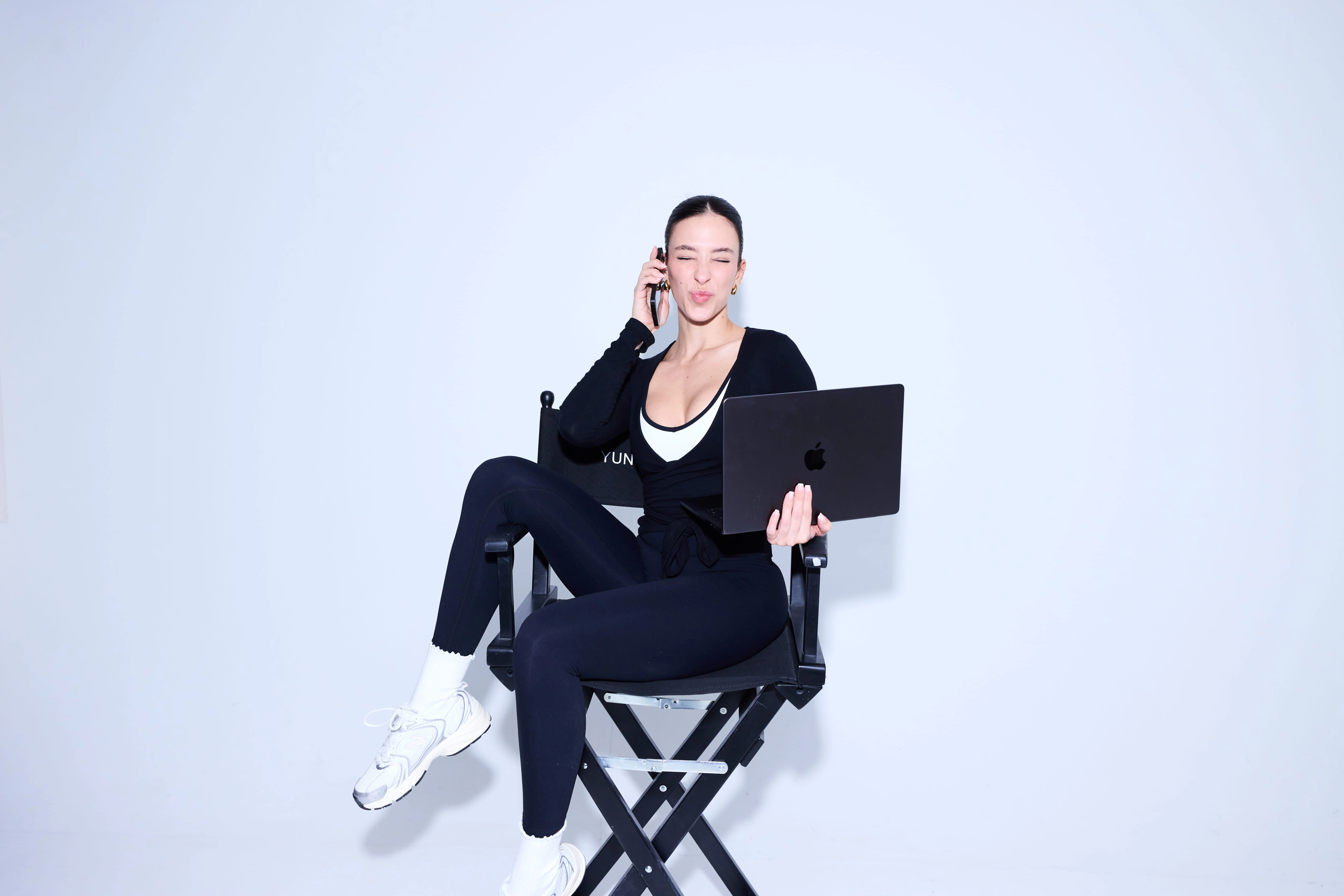 Young woman sitting cross-legged on a director's chair holding a laptop in one hand and a phone to her ear with a playful expression.