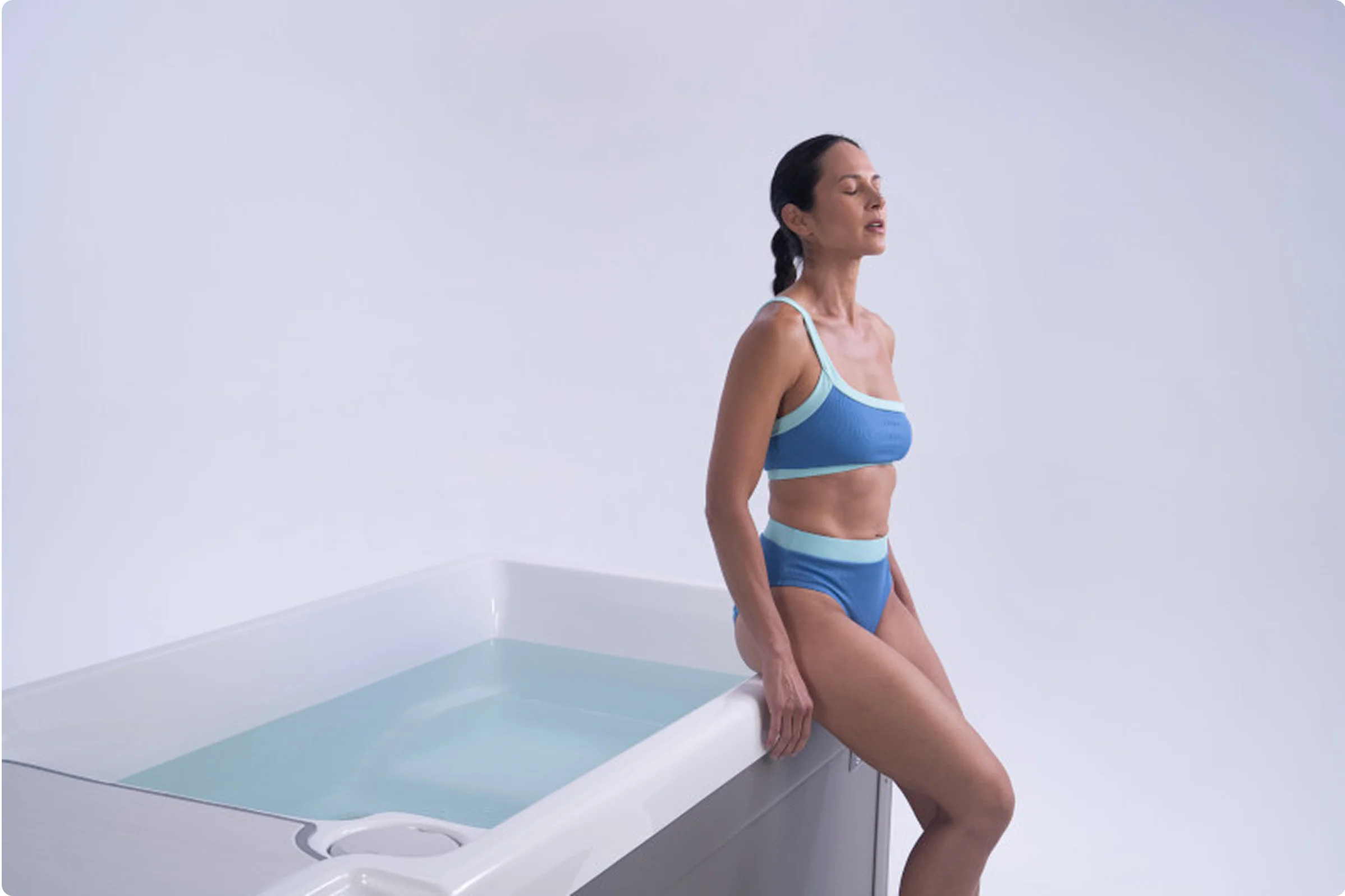 Eine Frau in einem blauen Badeanzug sitzt entspannt auf dem Rand eines Whirlpools, umgeben von einem minimalistischen, hellen Ambiente. Die geschlossene Augenhaltung und die ruhige Umgebung vermitteln ein Gefühl von Frieden und Erholung im Spa.