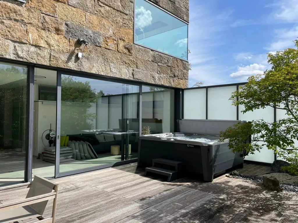 Modernes Haus mit Whirlpool auf Holzterrasse, umgeben von Glaswänden
