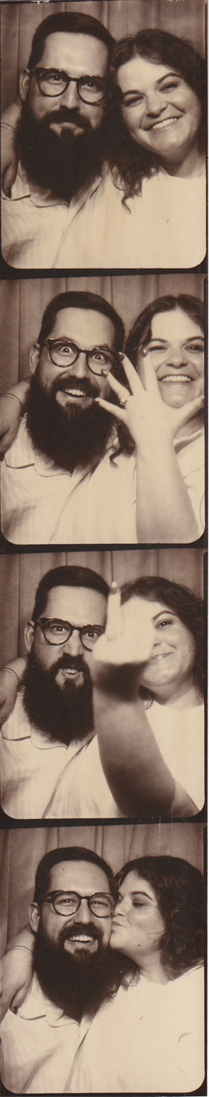 Photo booth strip showing a couple with the man wearing glasses and a beard, smiling and making playful faces, including the woman showing her hand with an engagement ring, the man showing his middle finger, and ending with the woman kissing the man on the cheek.