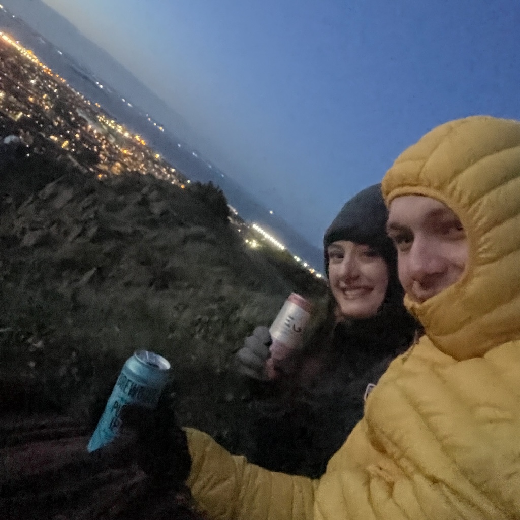 Two people dressed in warm jackets holding canned drinks with a city lit up in the background during dusk.