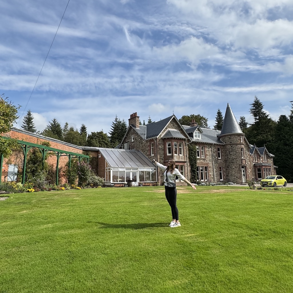 Woman standing with arms outstretched on green lawn in front of a large stone house with a conical turret and attached greenhouse.