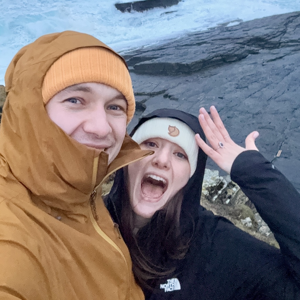 Smiling man in brown jacket and orange beanie with woman in black jacket and cream beanie showing a ring by the ocean rocks.