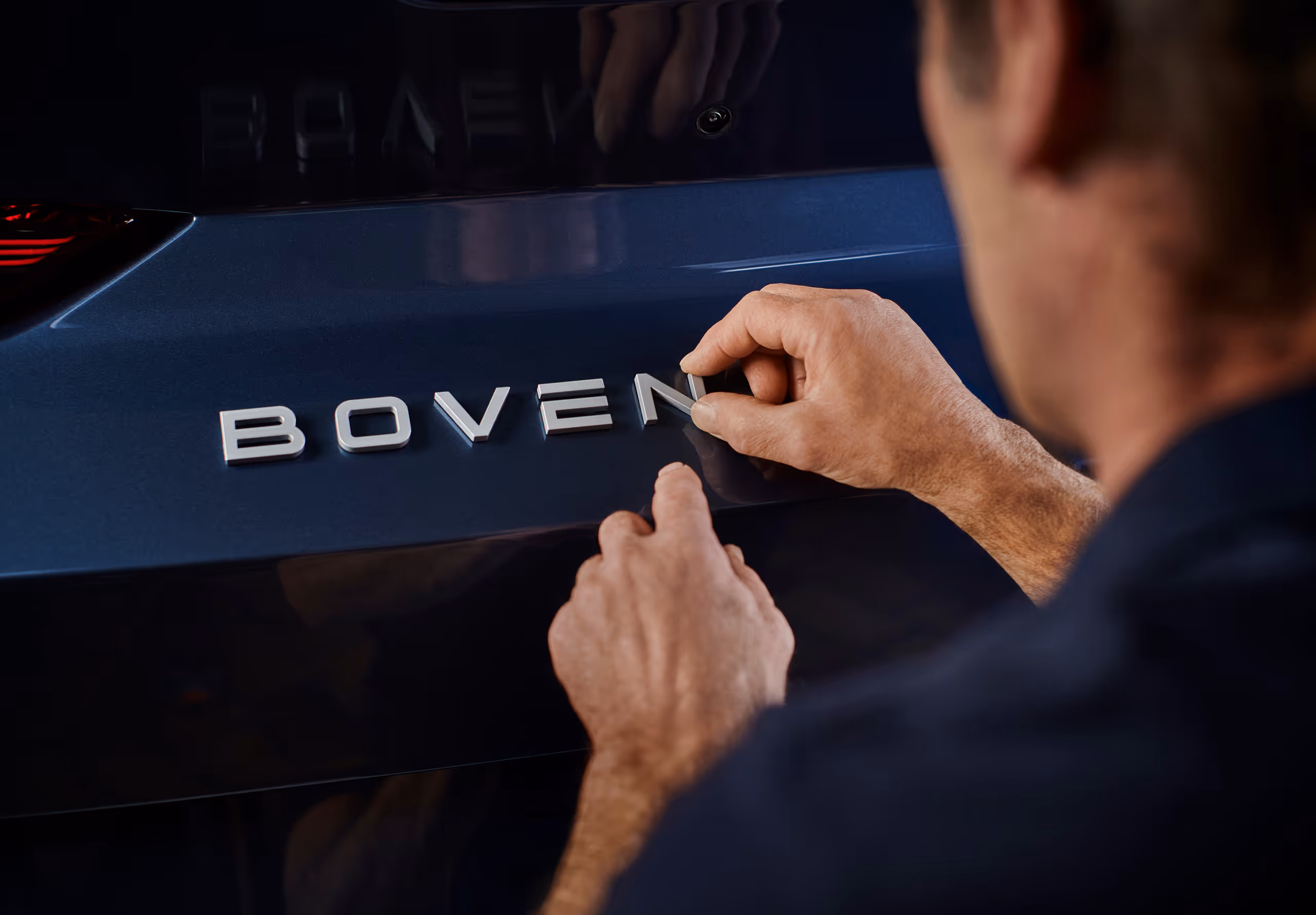 Person applying silver letters spelling BOVEN on the dark blue surface of a vehicle.