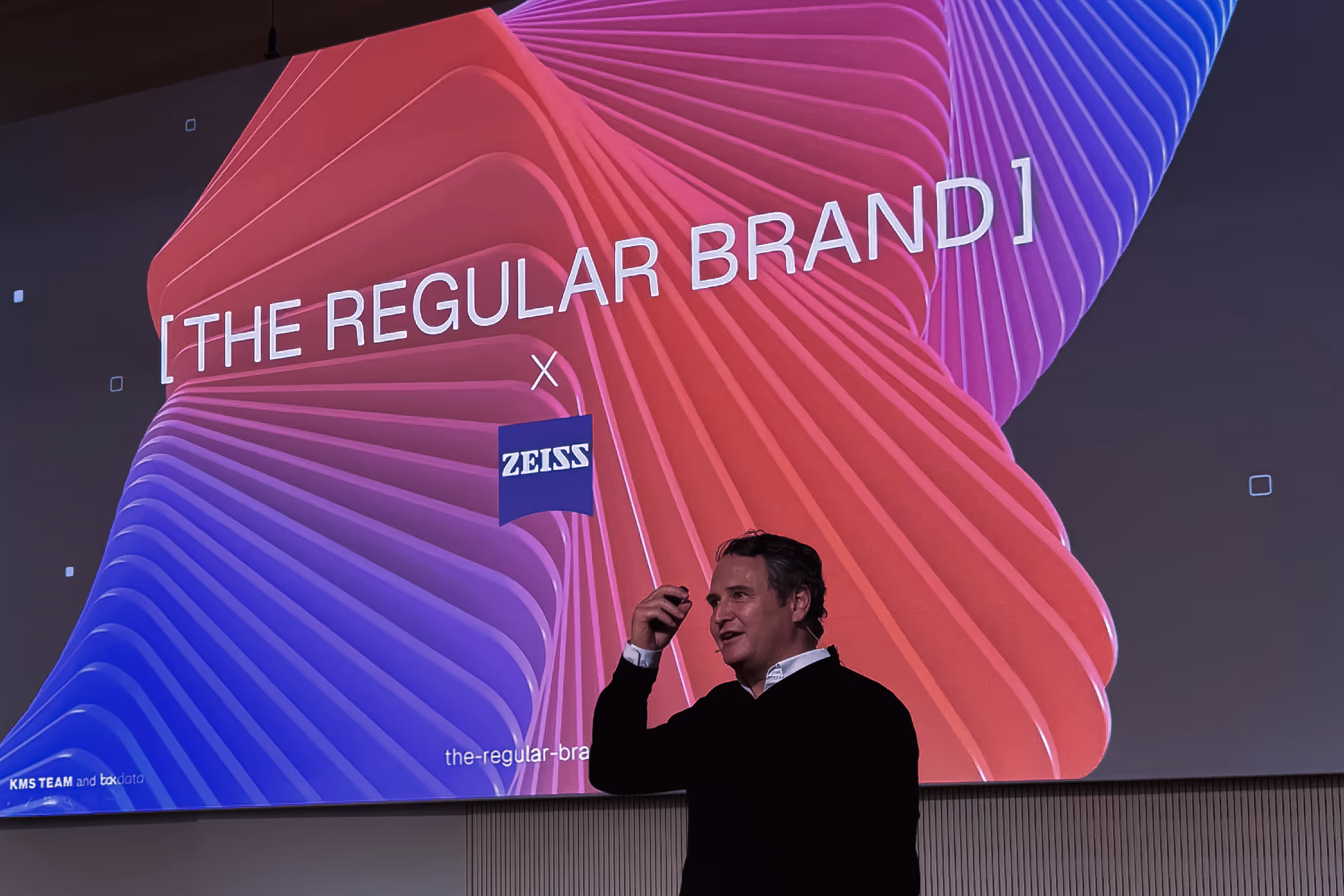 Man in black sweater giving a presentation in front of a screen with colorful abstract shapes and text 'THE REGULAR BRAND' and 'ZEISS'.