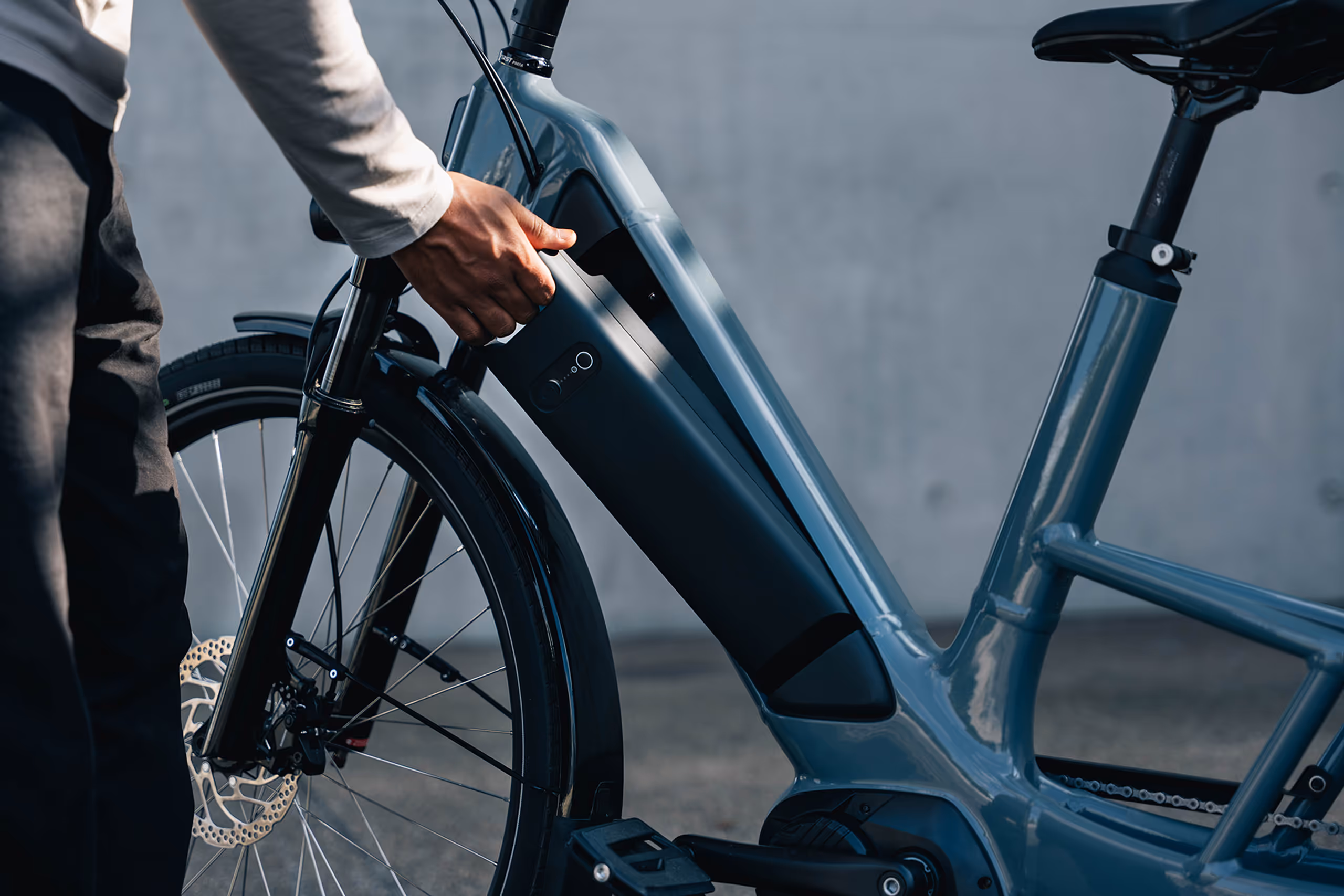 Person holding the battery of a blue electric bicycle near the front wheel and seat.