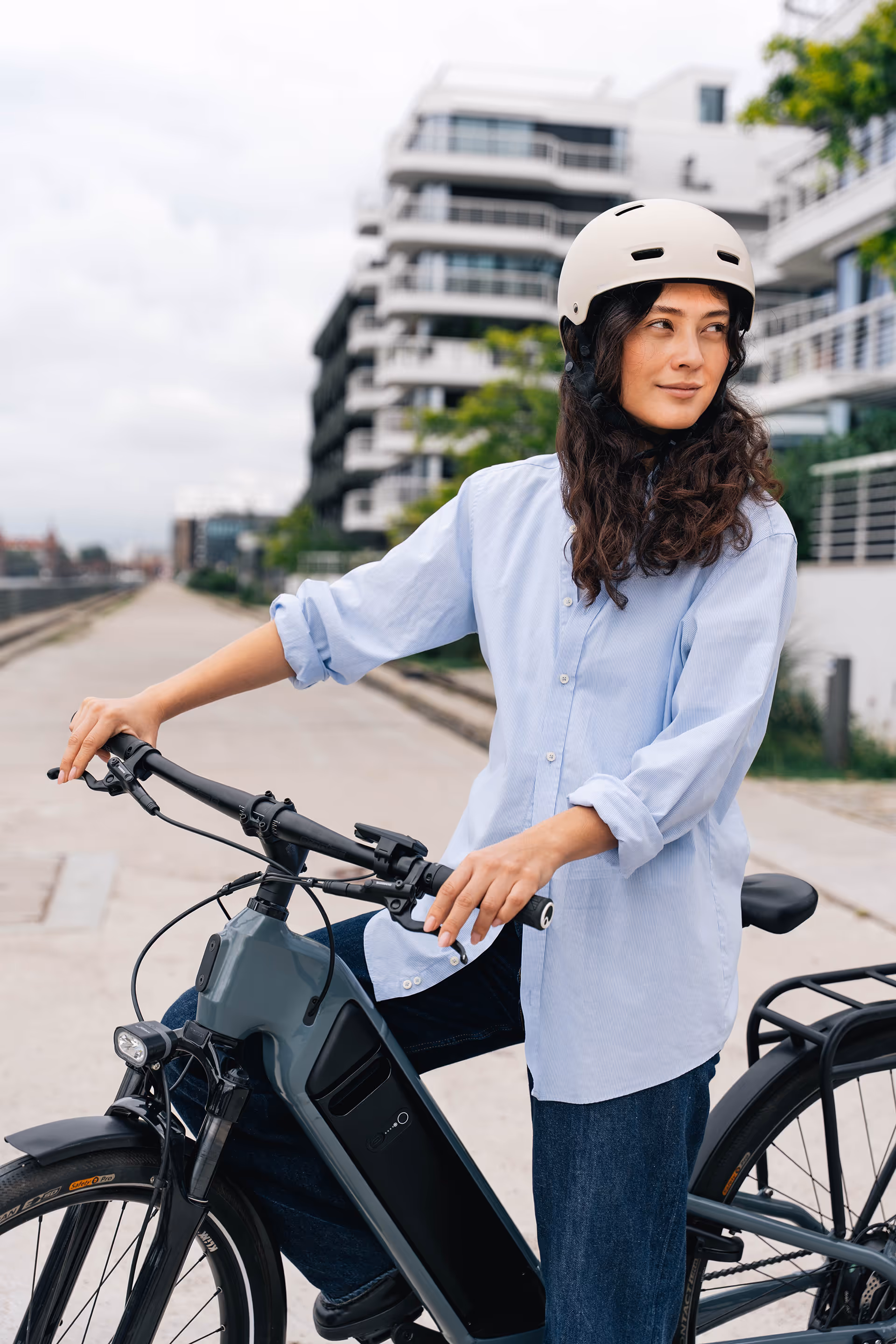 Woman wearing a white helmet and light blue shirt sitting on a black electric bicycle on a city sidewalk.