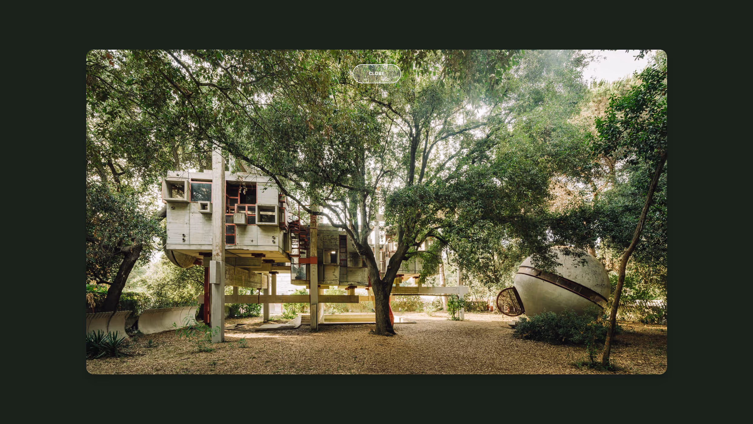 Modern house elevated on stilts surrounded by trees with a spherical concrete structure nearby in a wooded area.