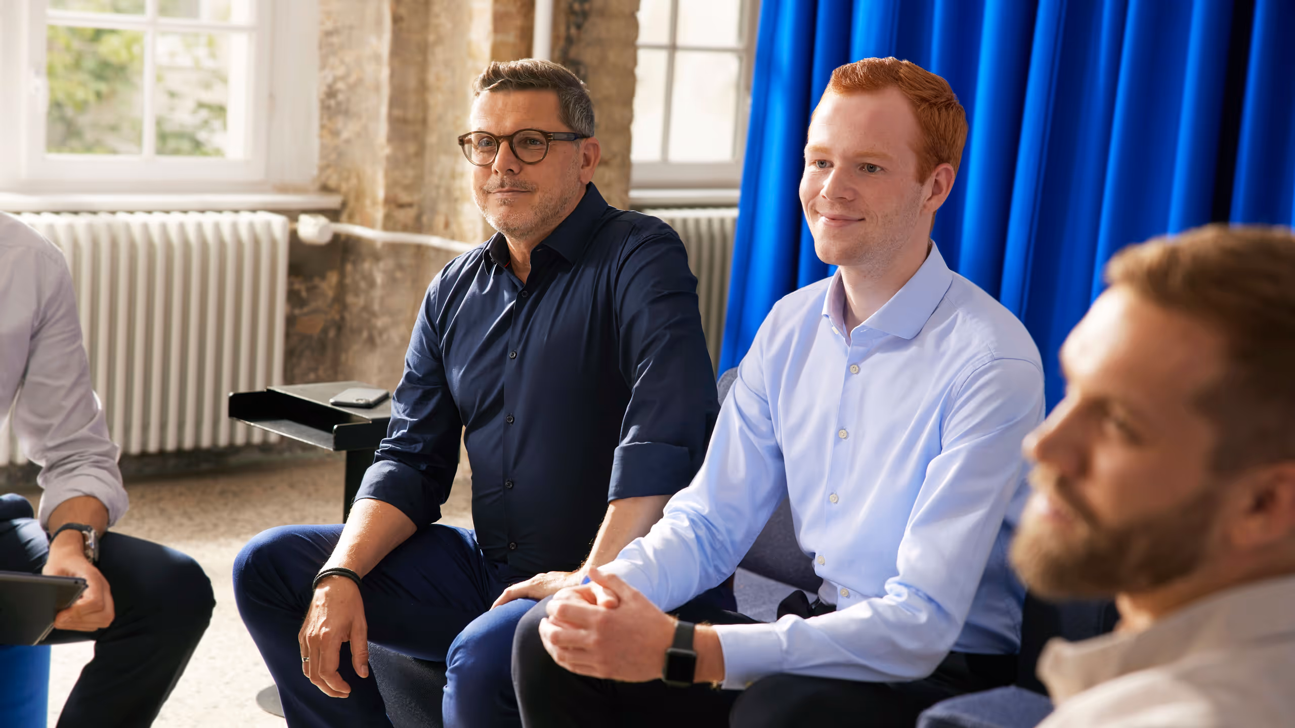 Four men sitting and attentively listening during a casual indoor meeting with blue curtains and windows in the background.