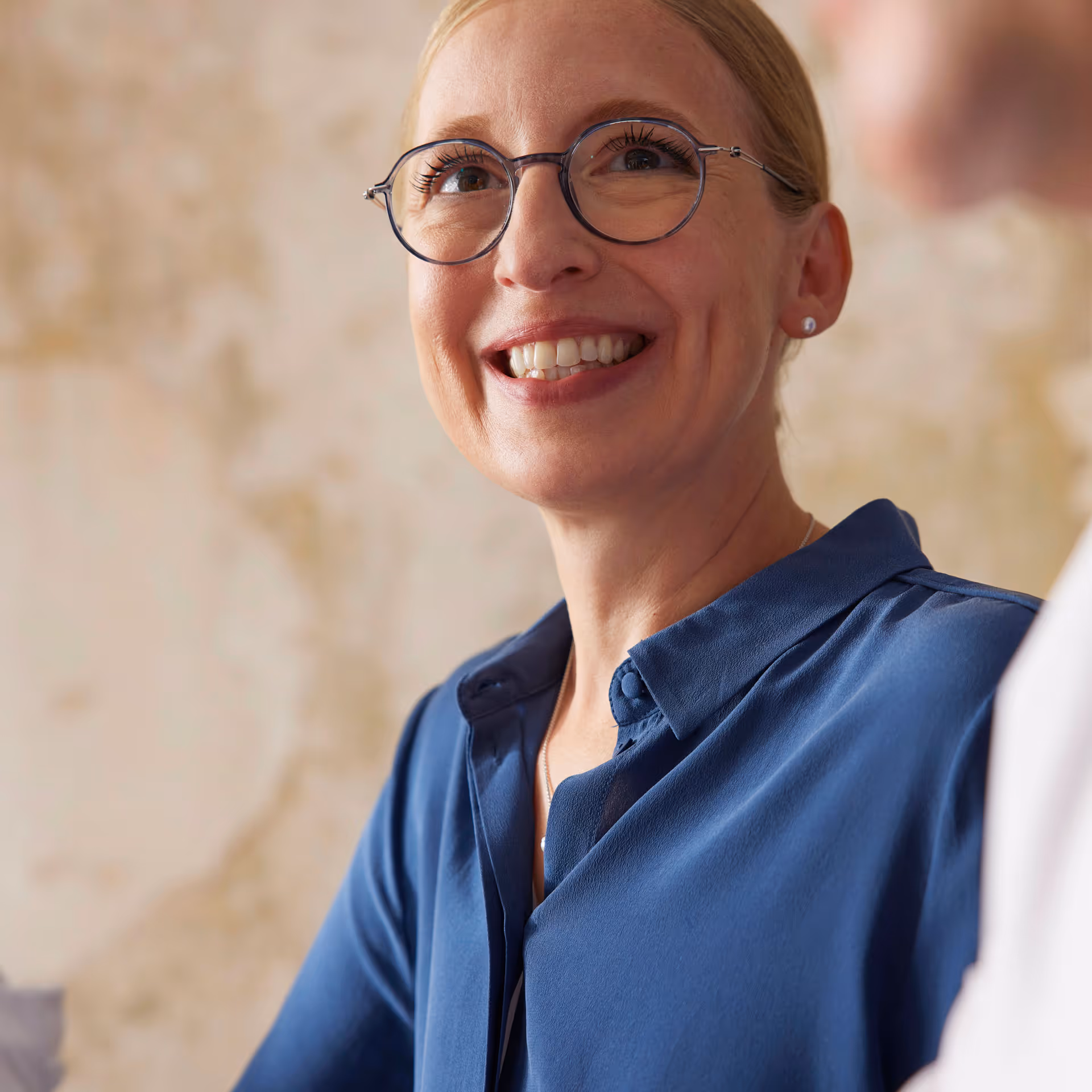Smiling woman with glasses wearing a blue shirt in conversation, with a blurred background.