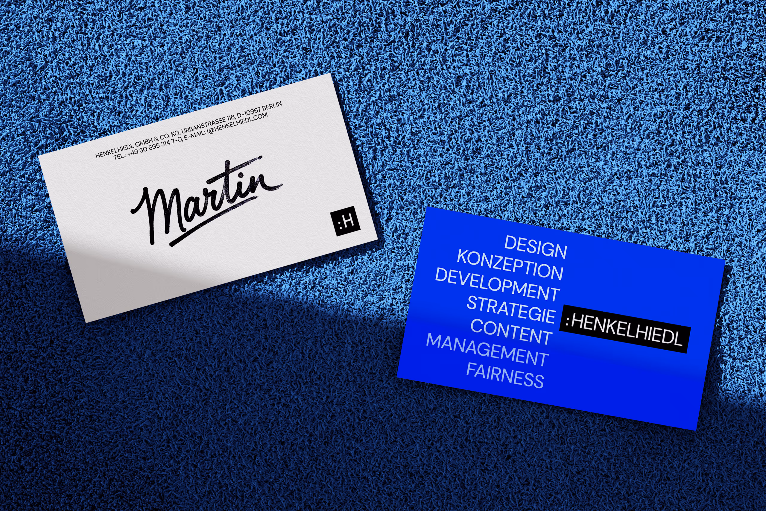 Two business cards on textured blue surface; one white with black handwritten name 'Martin' and contact info, one blue with white text listing services and brand name Henkelhiedl.
