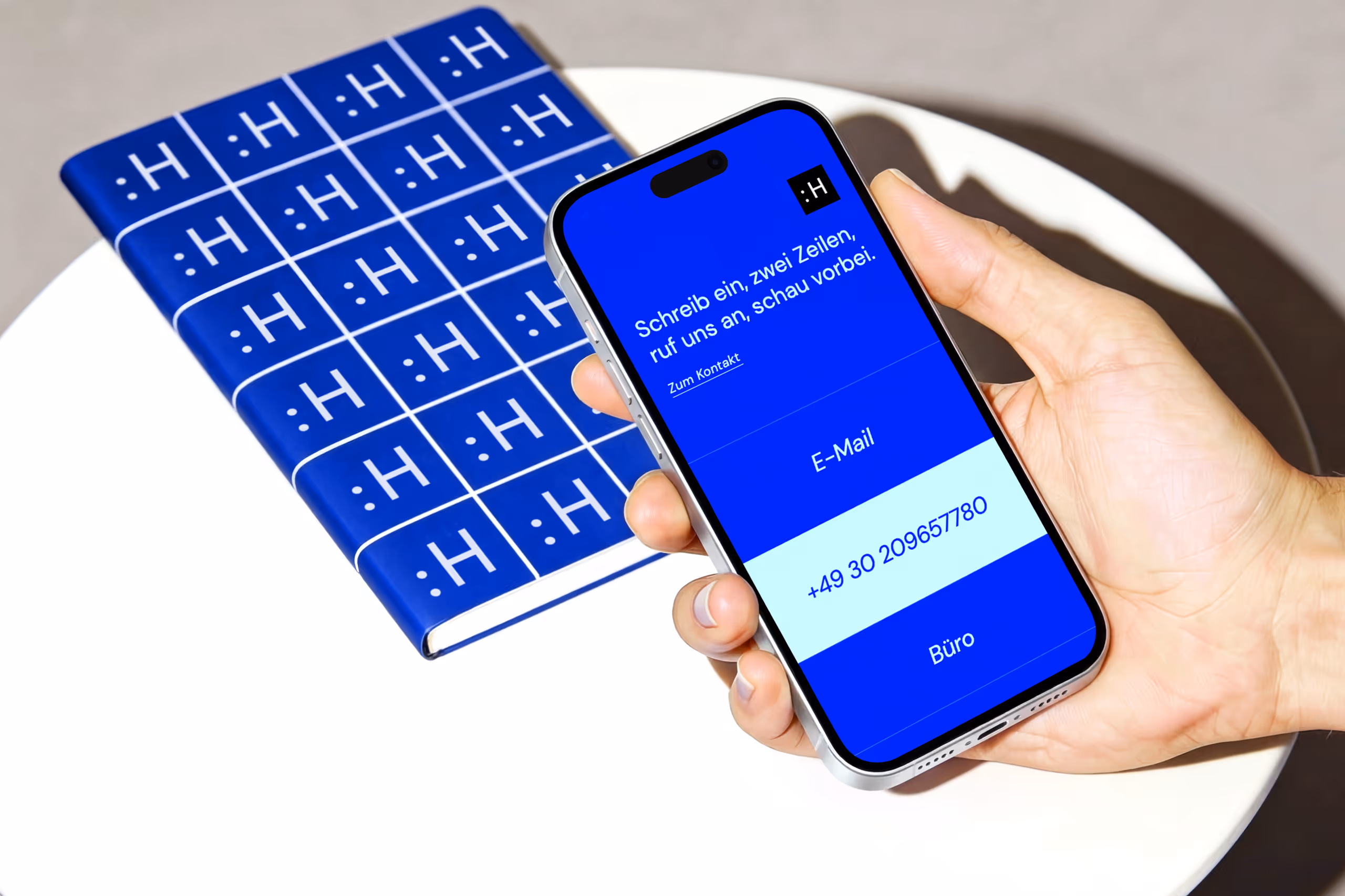 Hand holding a smartphone displaying a blue screen with German text and a phone number, next to a blue notebook with a repeating white ':H' pattern on a white round table.