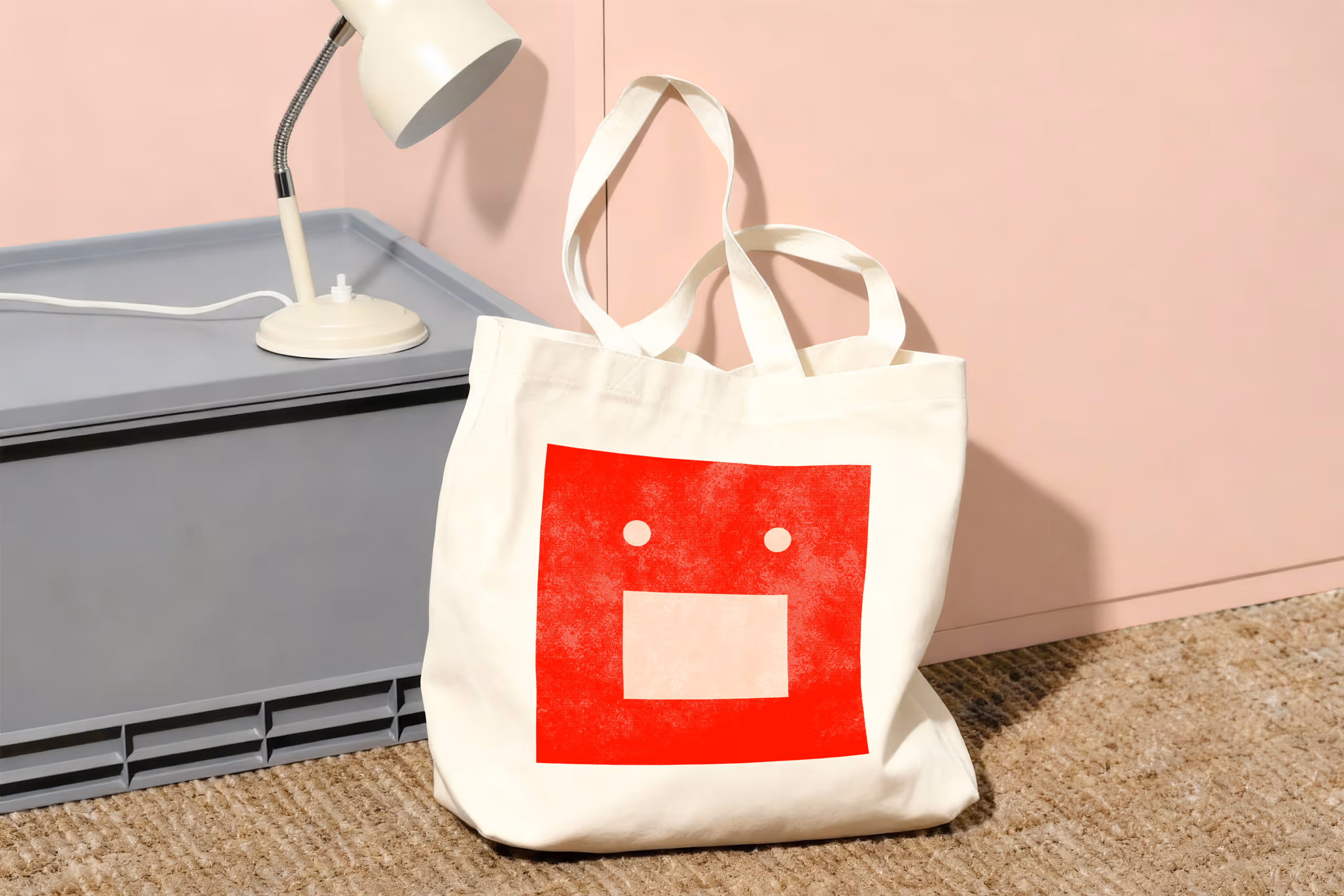 White tote bag with a red square design featuring two white dots and a larger white rectangle, placed on a textured brown floor near a gray box and a white lamp.
