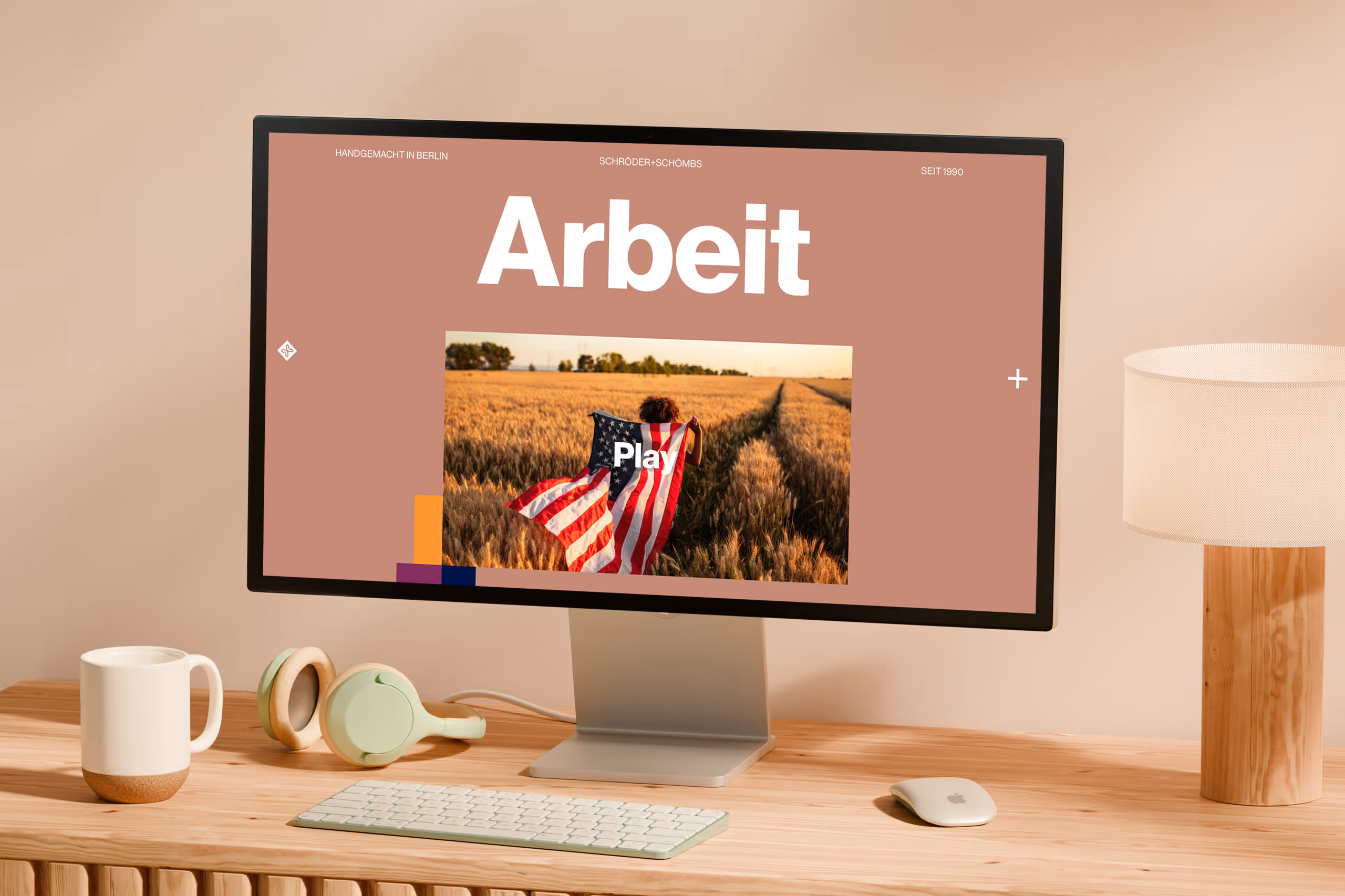 Desktop computer on wooden desk displaying a website with the word 'Arbeit' and a play button over a field image, surrounded by headphones, a mug, and a lamp.