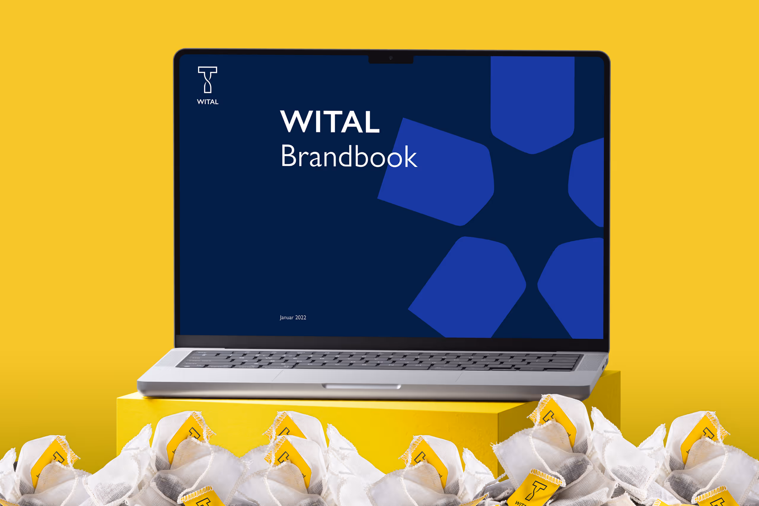 Laptop displaying a WITAL Brandbook cover on a blue screen, placed on a yellow box with tea bags labeled with the WITAL logo in front.