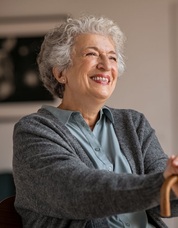 Smiling elderly woman with curly gray hair wearing a gray cardigan and light blue shirt, holding a wooden cane.