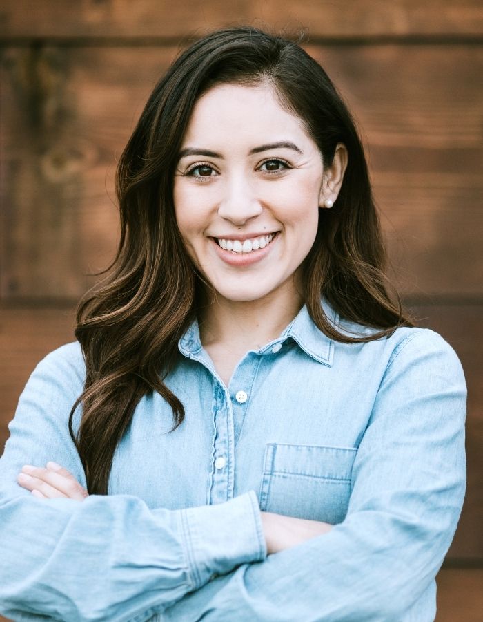 Smiling woman with long dark hair wearing a light blue denim shirt, standing with arms crossed against a wooden background.