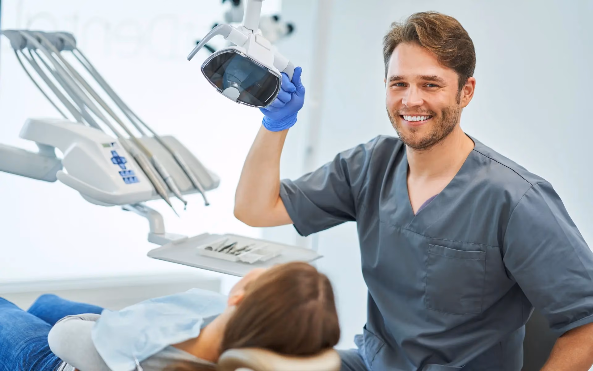 Male dentist in gray scrubs smiling and adjusting dental light while a patient reclines in the dentist chair.