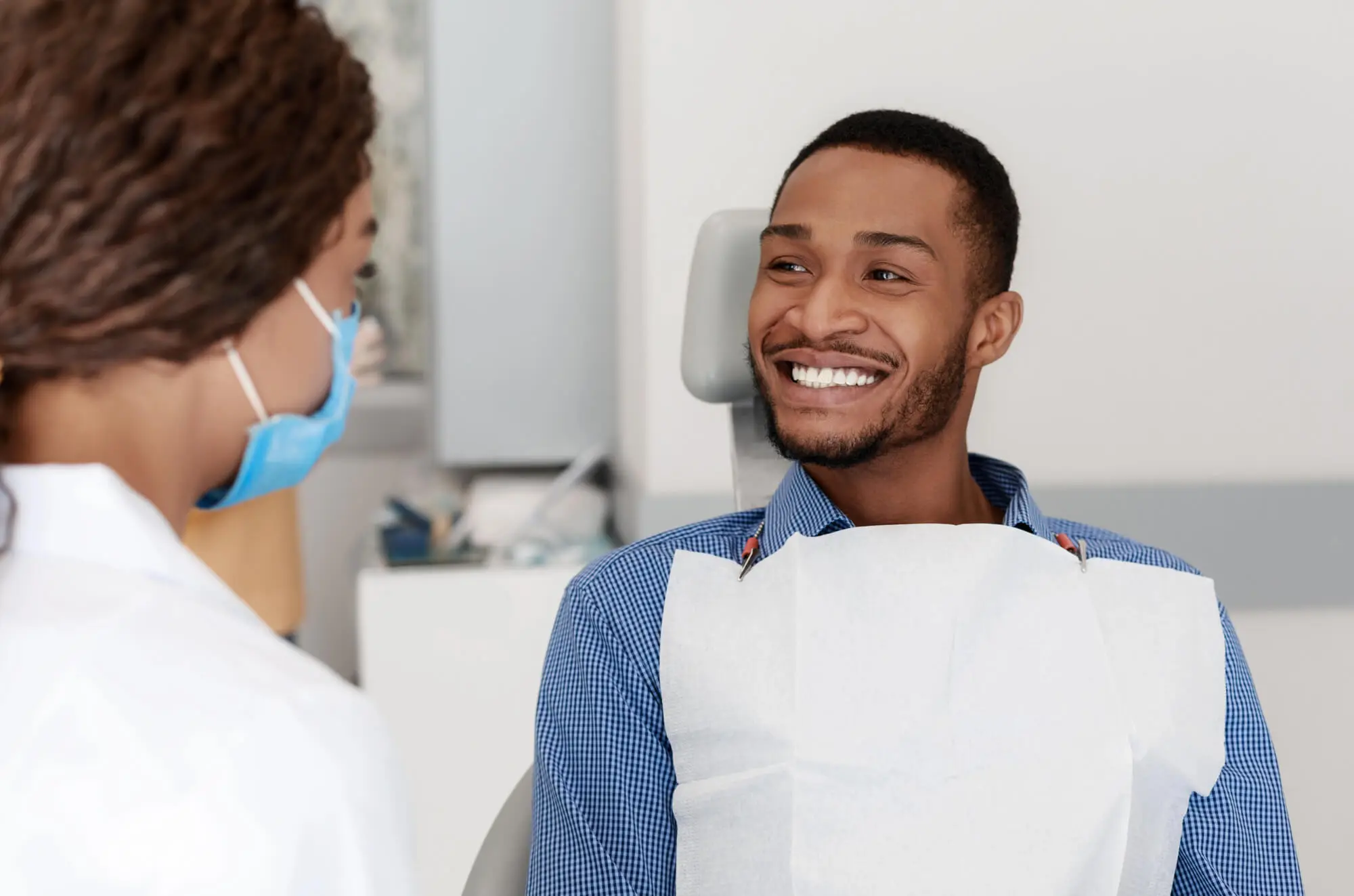 Smiling male patient in a dental chair talking with a female dentist wearing a blue face mask.