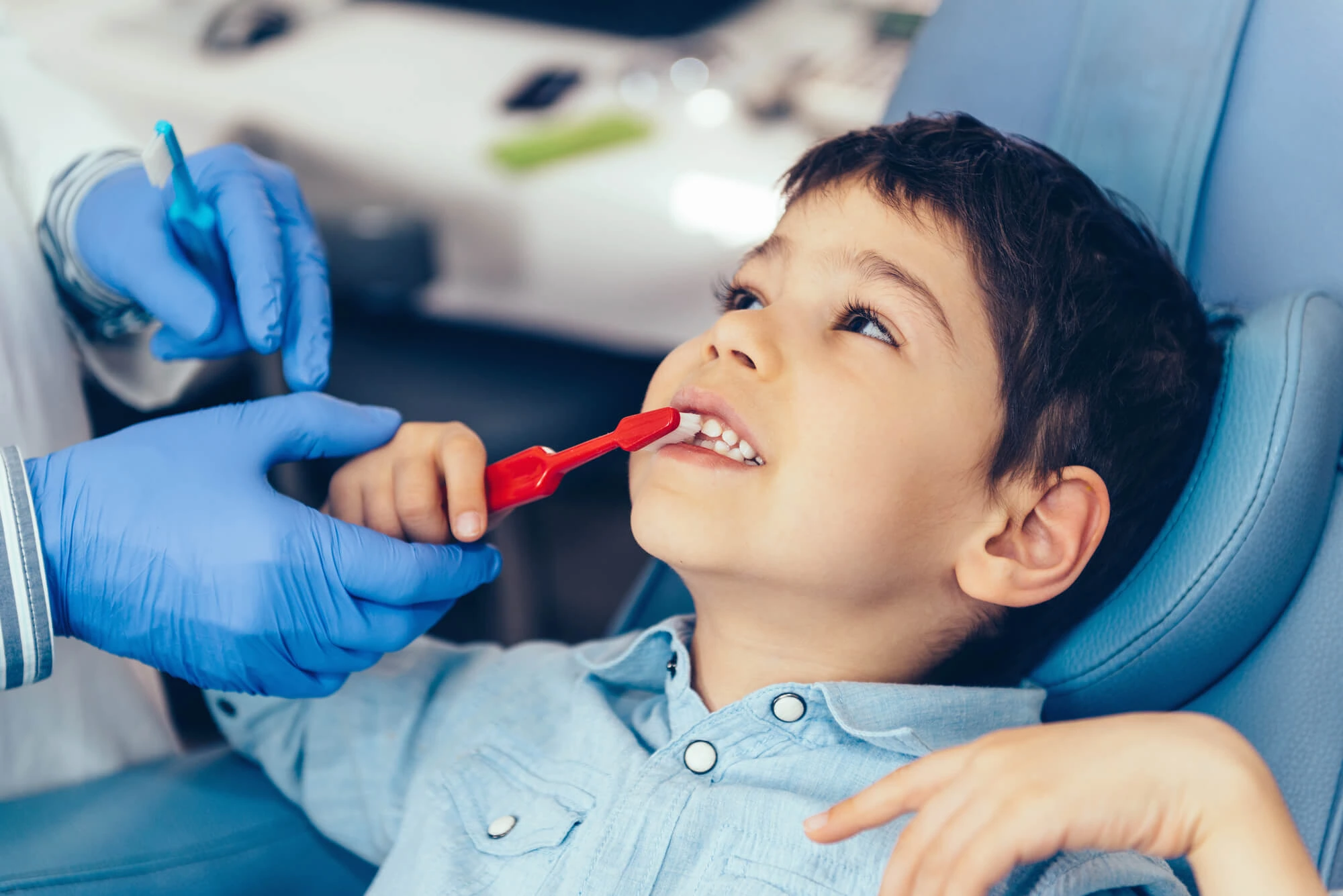 Dentist wearing blue gloves helping a young boy practice brushing teeth with a red toothbrush in a dental chair.