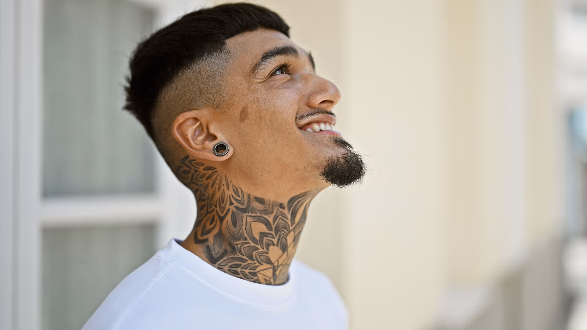Young man with a black floral neck tattoo looking upward and smiling, wearing a white shirt and black ear gauges.