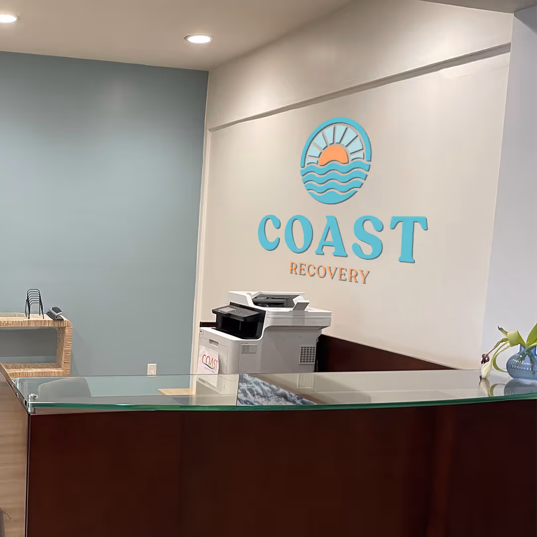 Reception area with a dark wood and glass desk, a white multifunction printer, and a wall sign reading 'COAST RECOVERY' with a sun and waves logo.