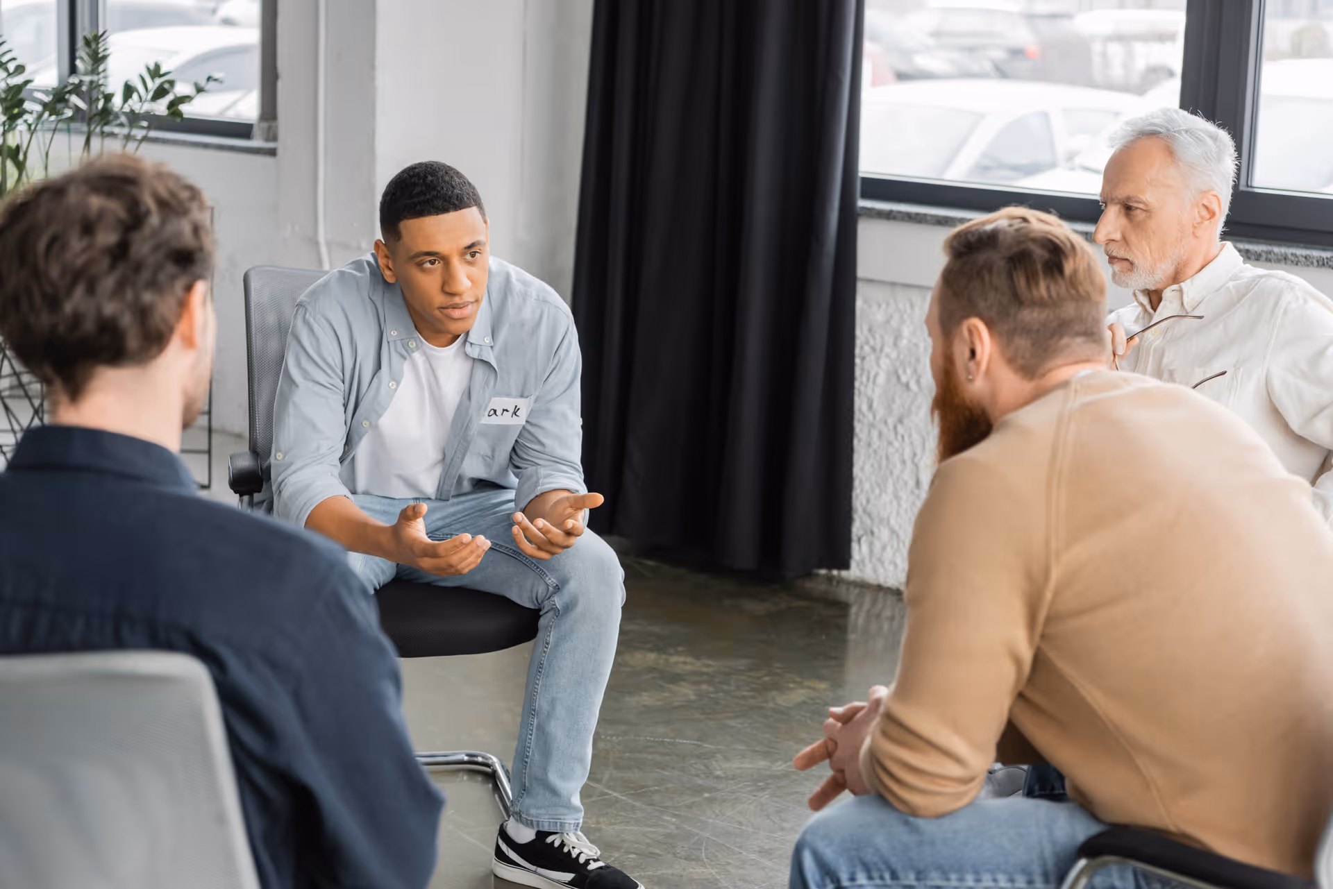 A young man is speaking seriously while three other men listen attentively in a group discussion setting.