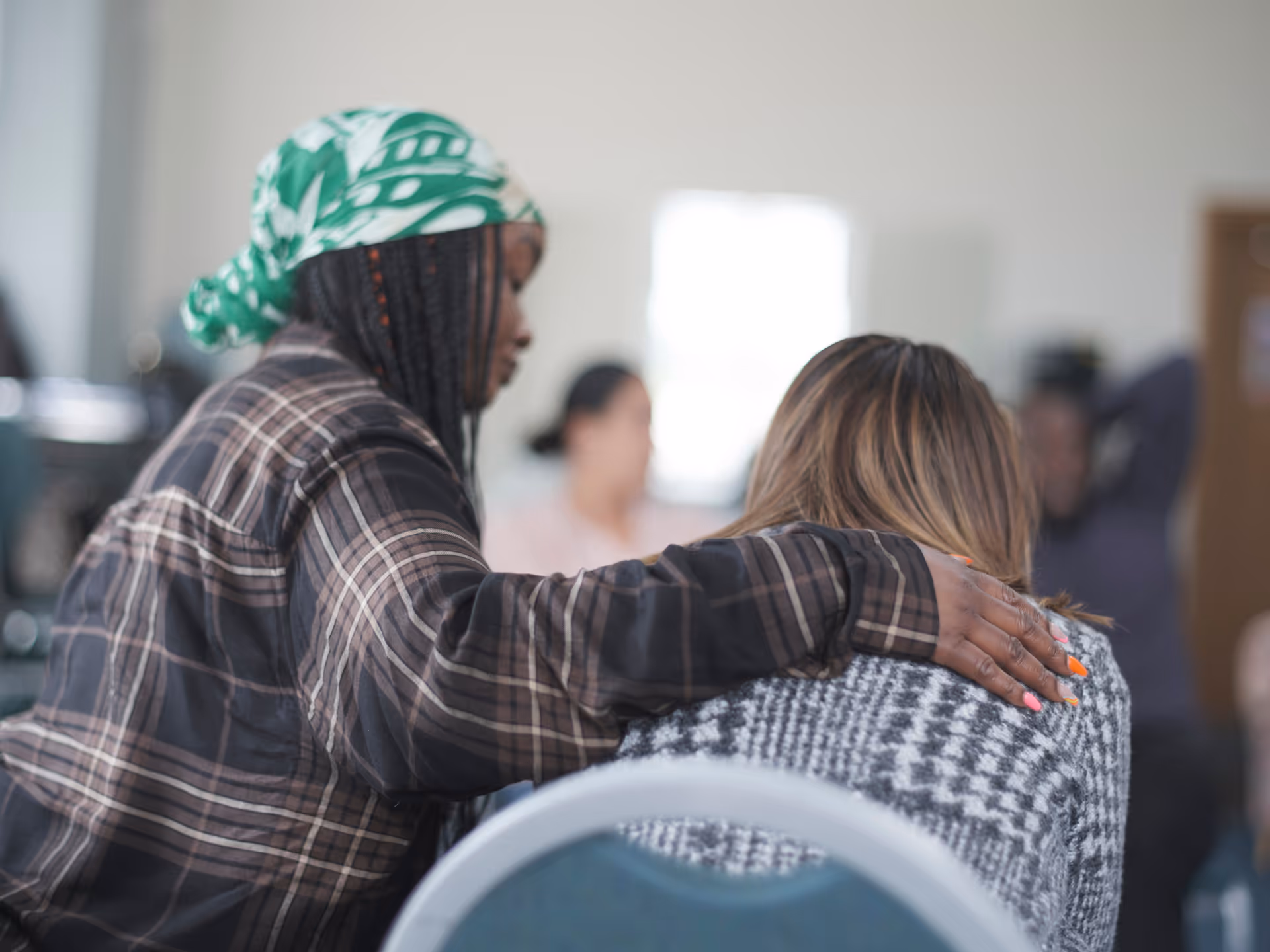 Person with a green and white headscarf and plaid shirt comforting another person by placing an arm around their shoulder.