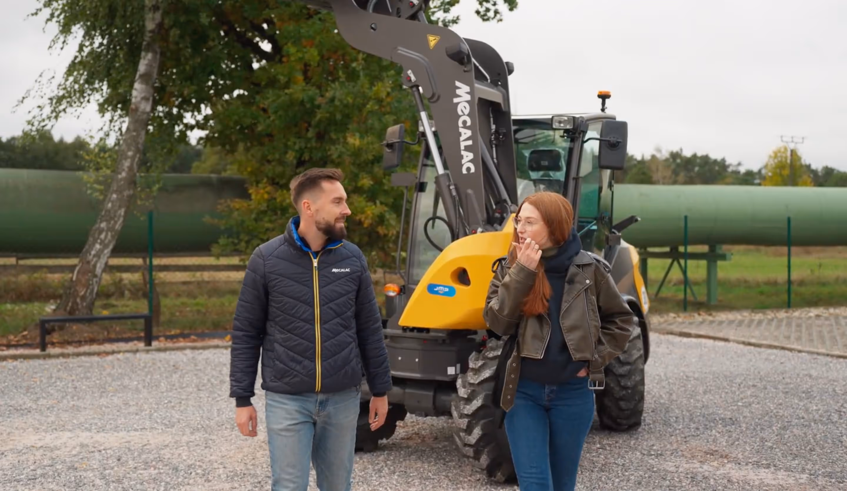 Muž a žena v jezdeckém oblečení kráčí vedle žlutého stavebního stroje Mecalac na štěrkové cestě s lesem v pozadí.
