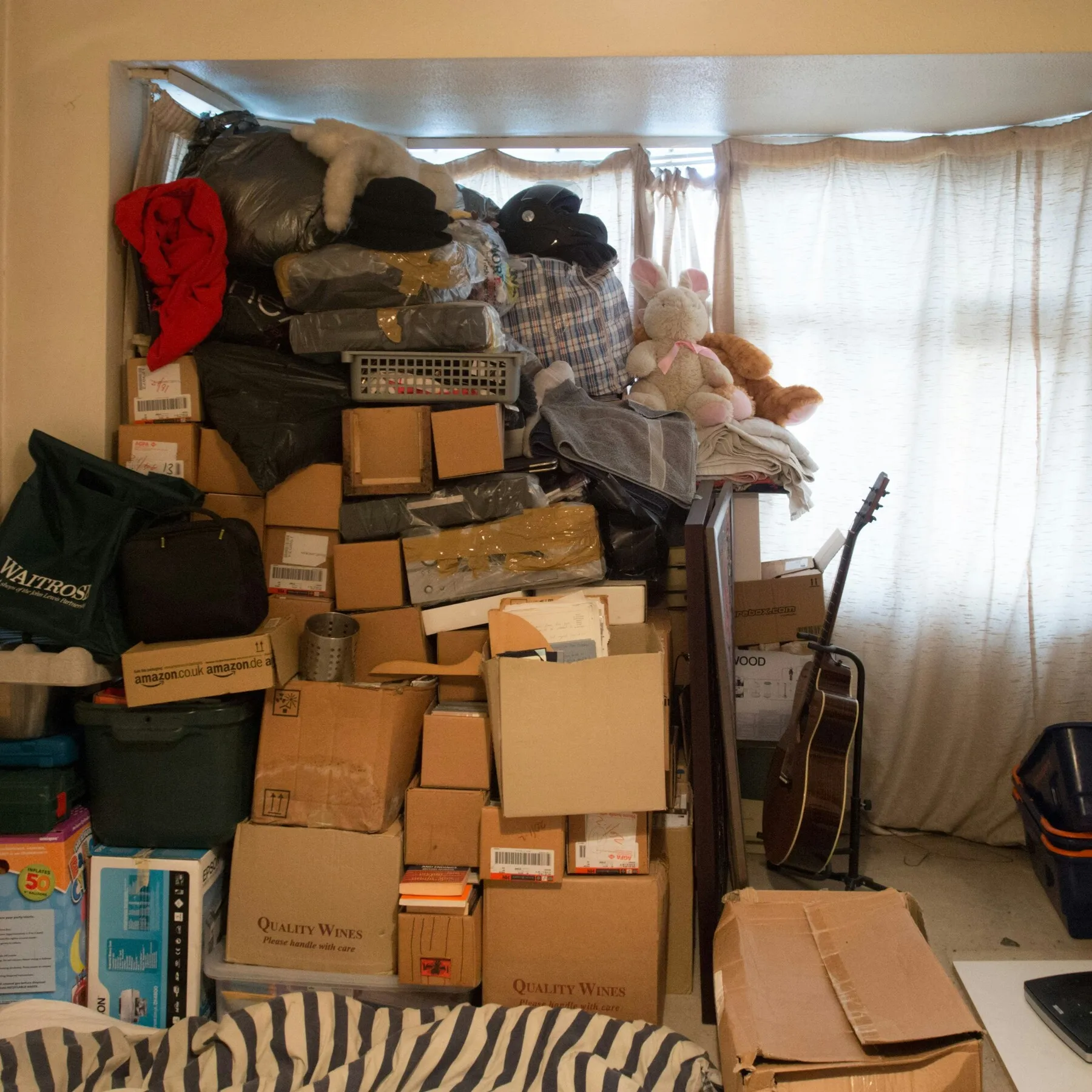 A cluttered room filled with stacked cardboard boxes, bags, and stuffed animals blocking a window — a common result of lacking extra space storage at home.