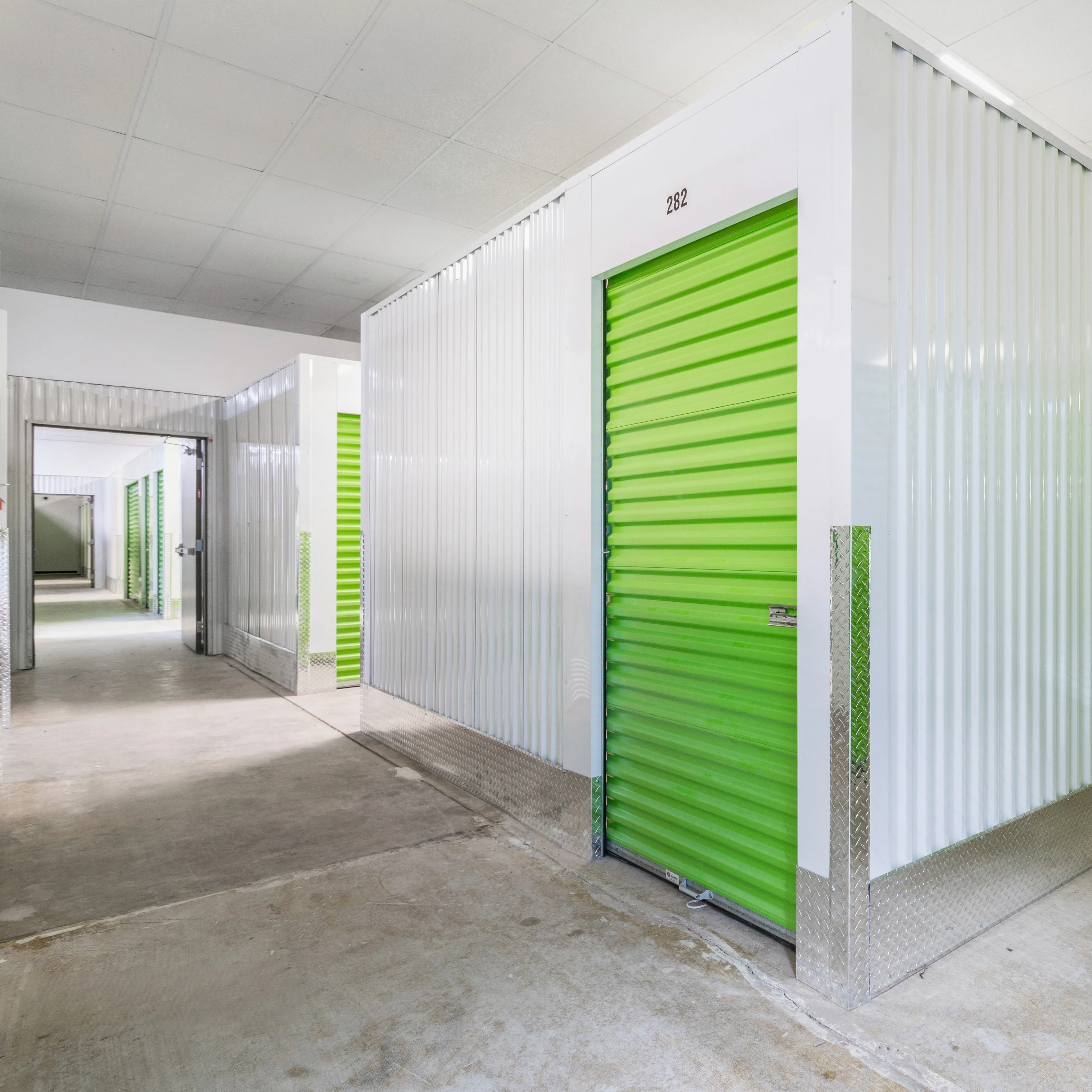Bright indoor climate-controlled storage facility with clean white walls and vibrant green roll-up unit doors, featuring polished concrete floors and secure access.