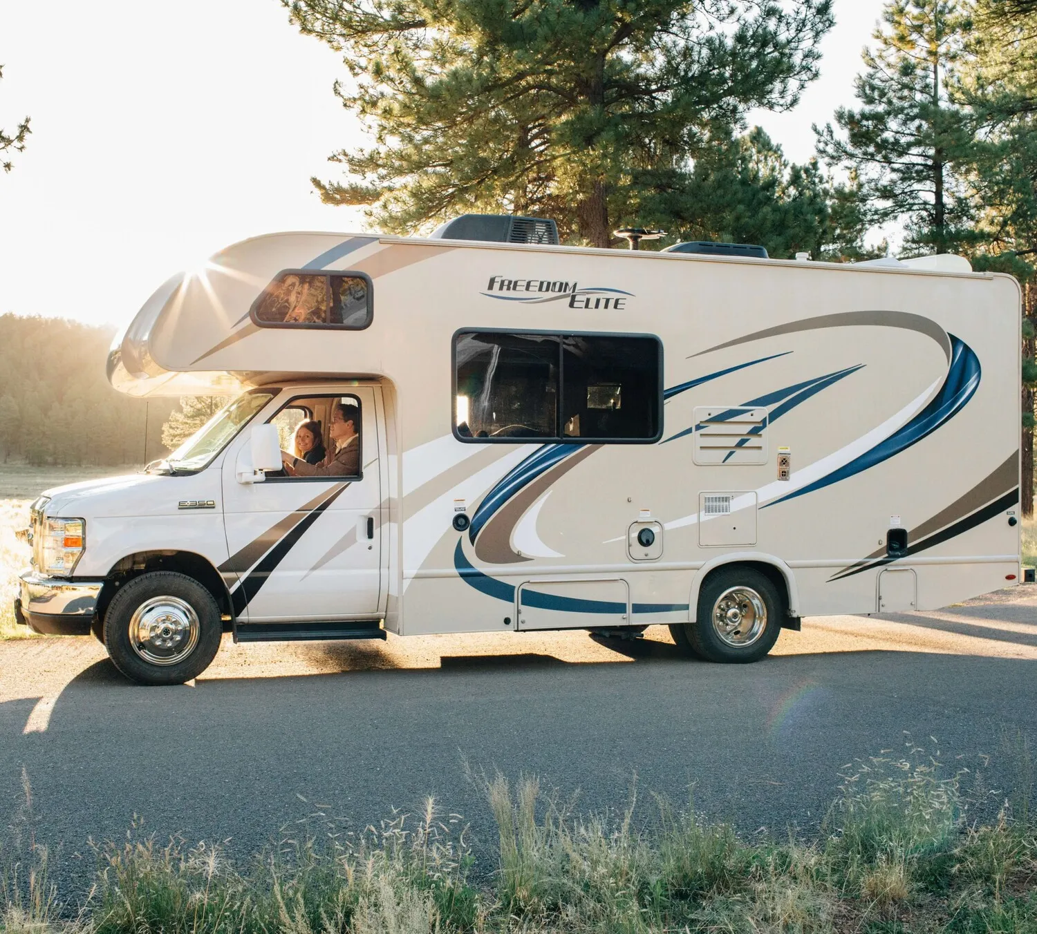 Class C RV parked near forest with couple inside enjoying the view through the front window