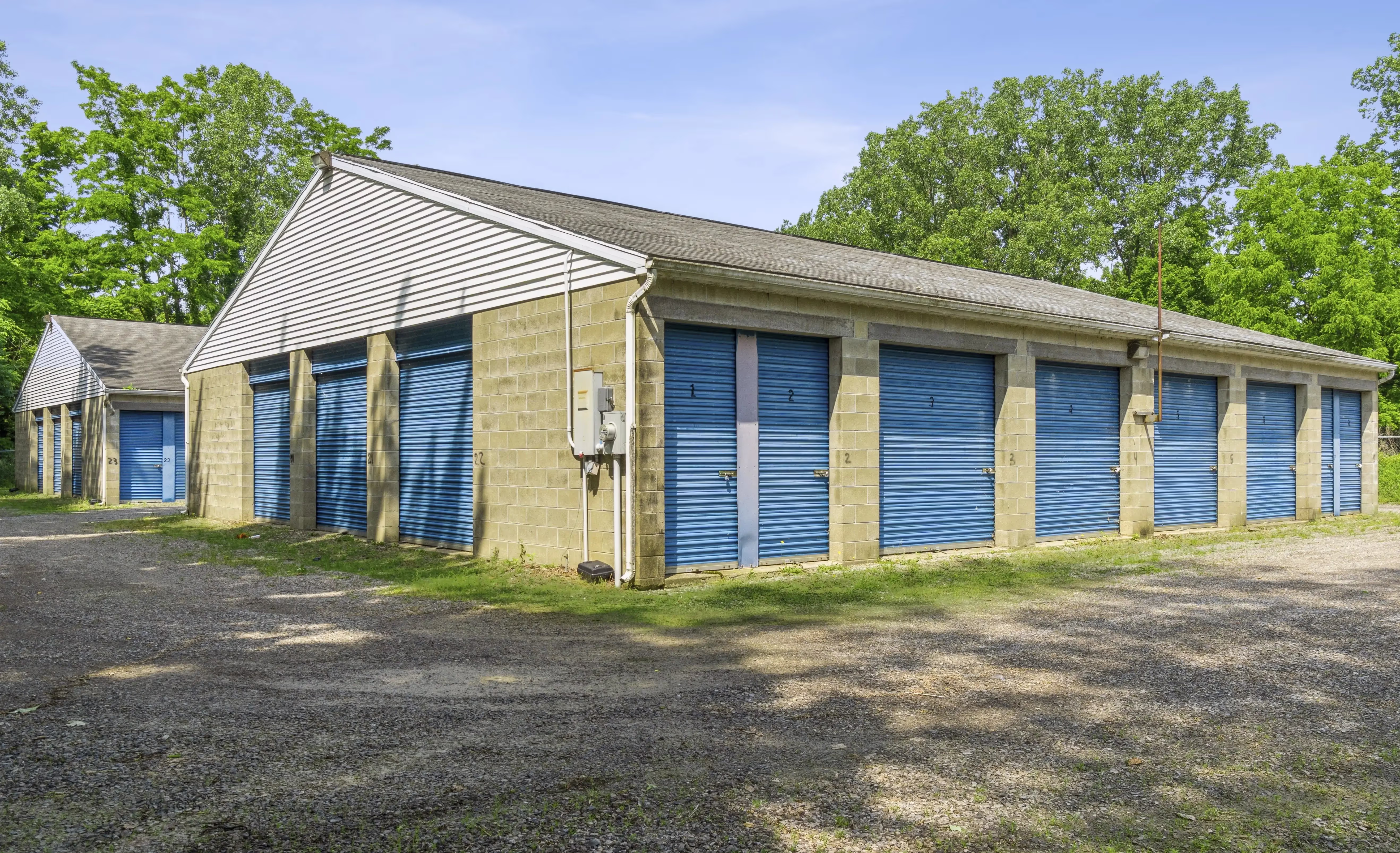 Row of drive up storage units with blue roll up doors surrounded by trees in Fairview PA
