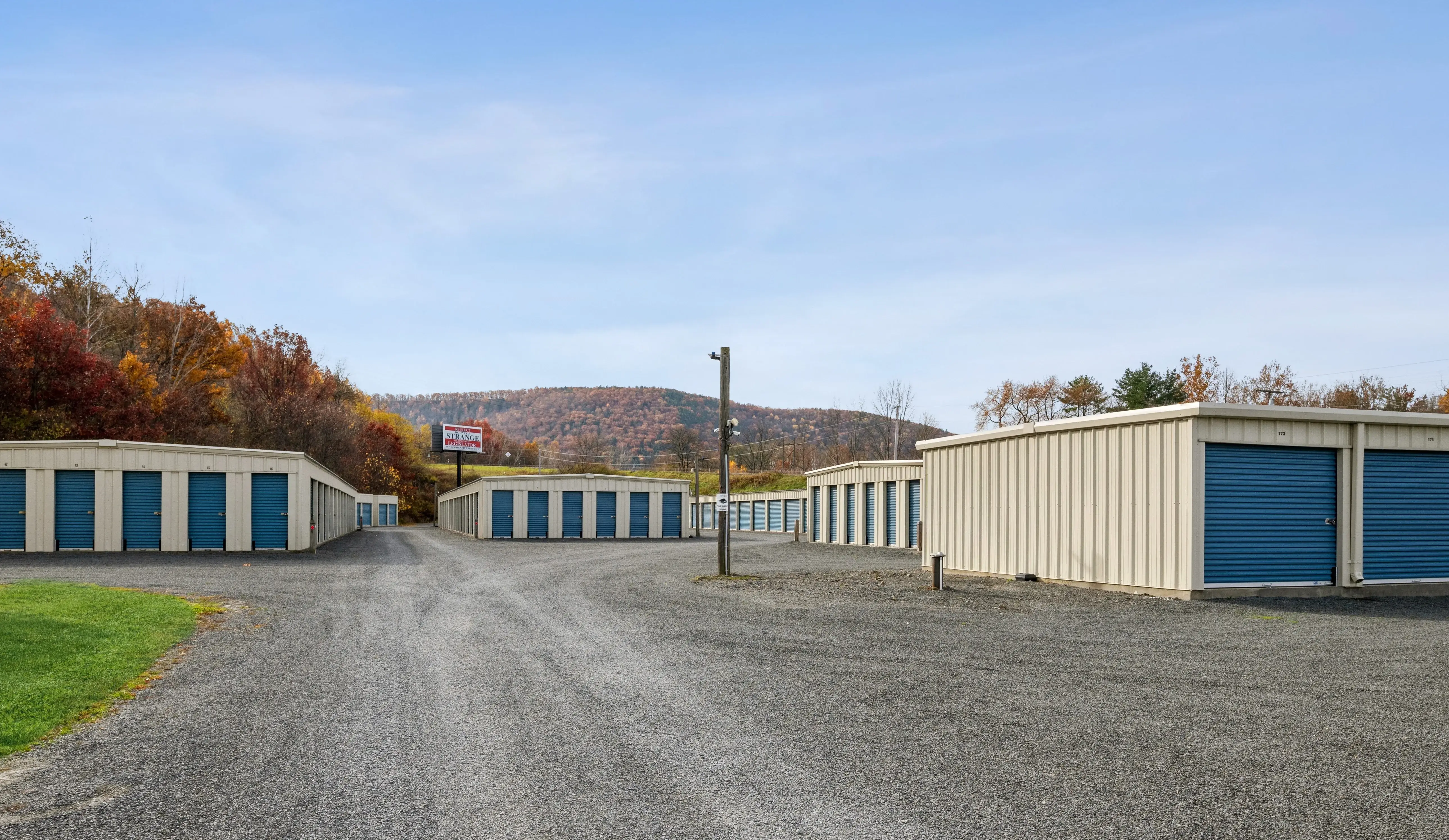 Drive up storage units with blue doors and gravel access lanes surrounded by fall trees in Elmira NY