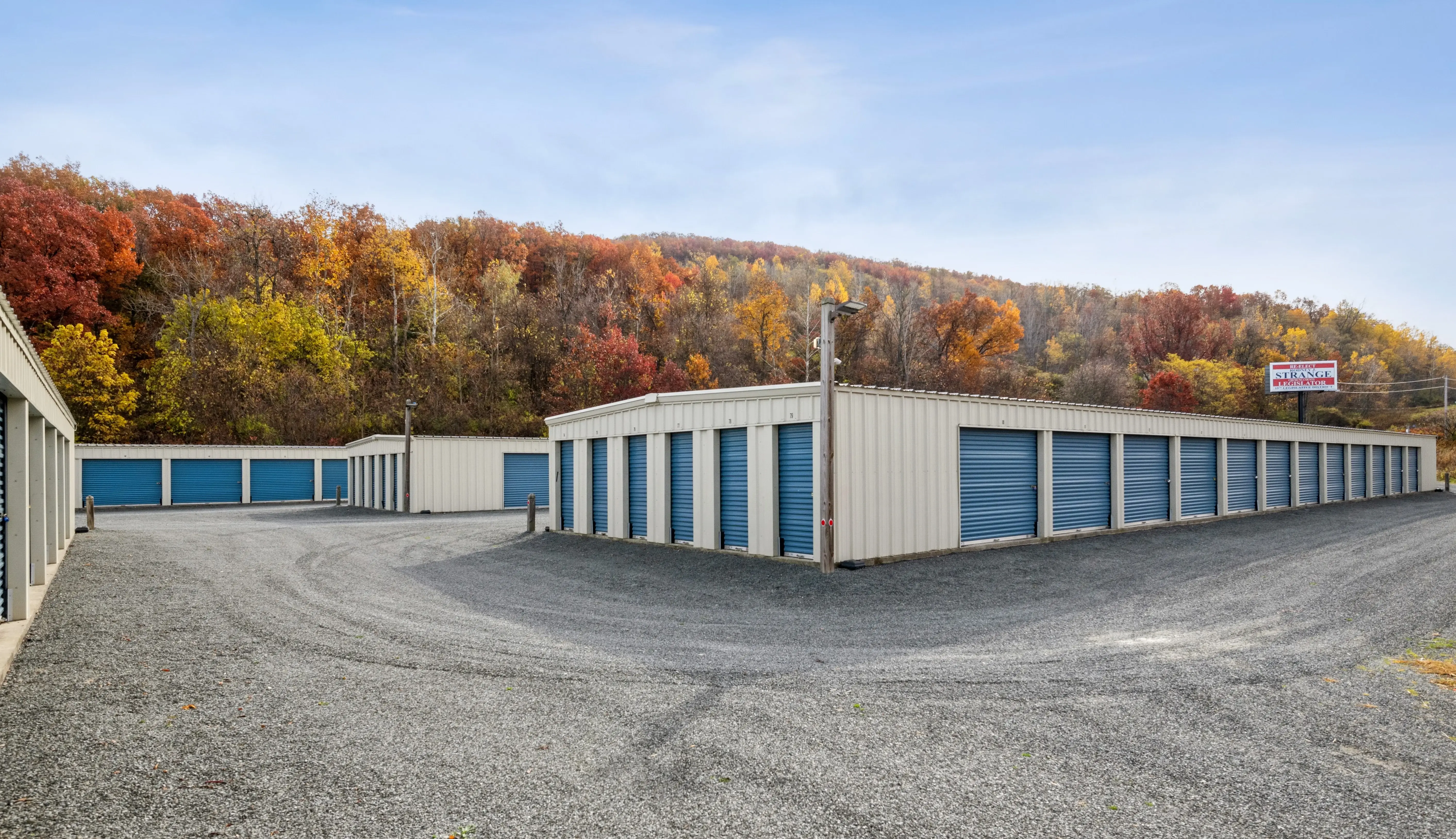Drive up storage units with blue doors and gravel access lanes surrounded by fall trees in Elmira NY