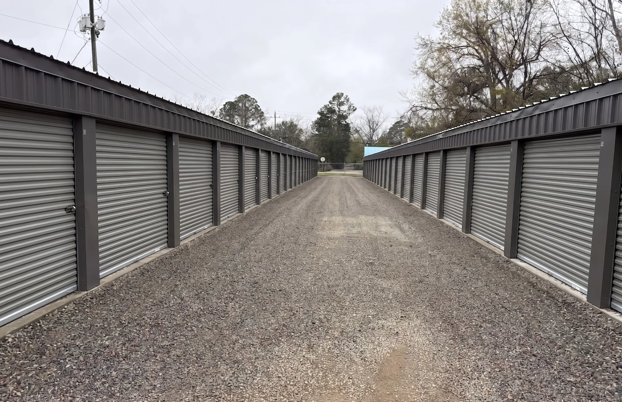Looking down a wide gravel path between two rows of outdoor storage units in Baxley, Georgia, showing the uniform grey roll up doors and metal building design