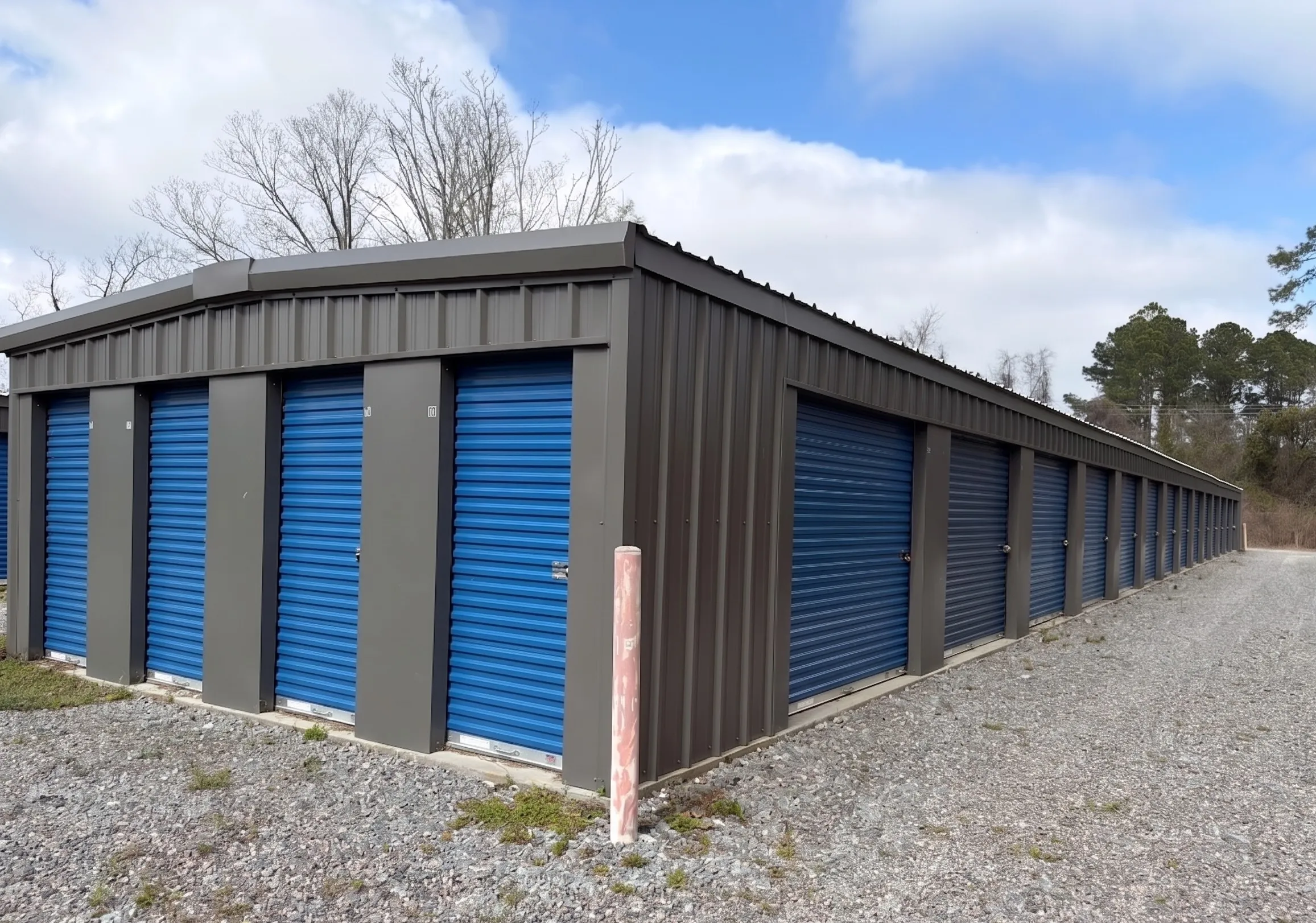 side-angle view of a grey metal storage unit building in Jesup, Georgia, showing a long row of secure blue roll up doors and a level gravel path for loading and unloading