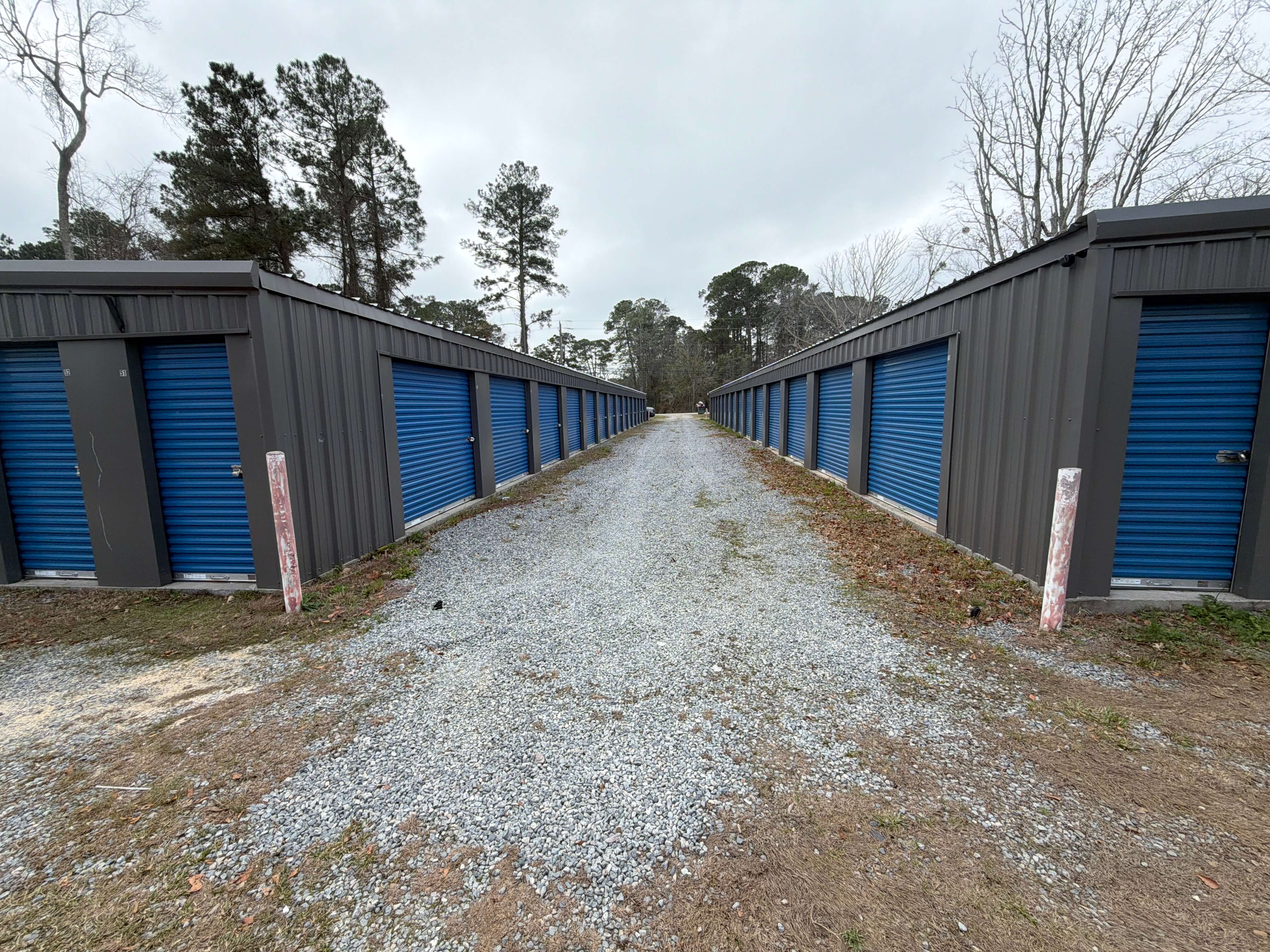 Looking down a gravel lane between two rows of storage units at 1160 W Cherry St, Jesup, GA 31545, with blue roll up doors on both sides and wide access for trucks