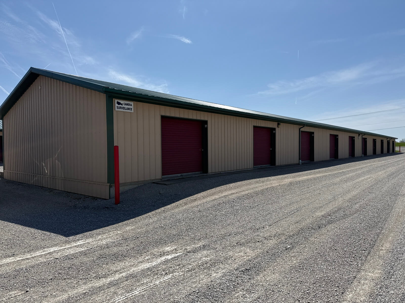 Self storage building exterior with numbered burgundy unit doors along a gravel drive at Bolt Storage Ossian, IN
